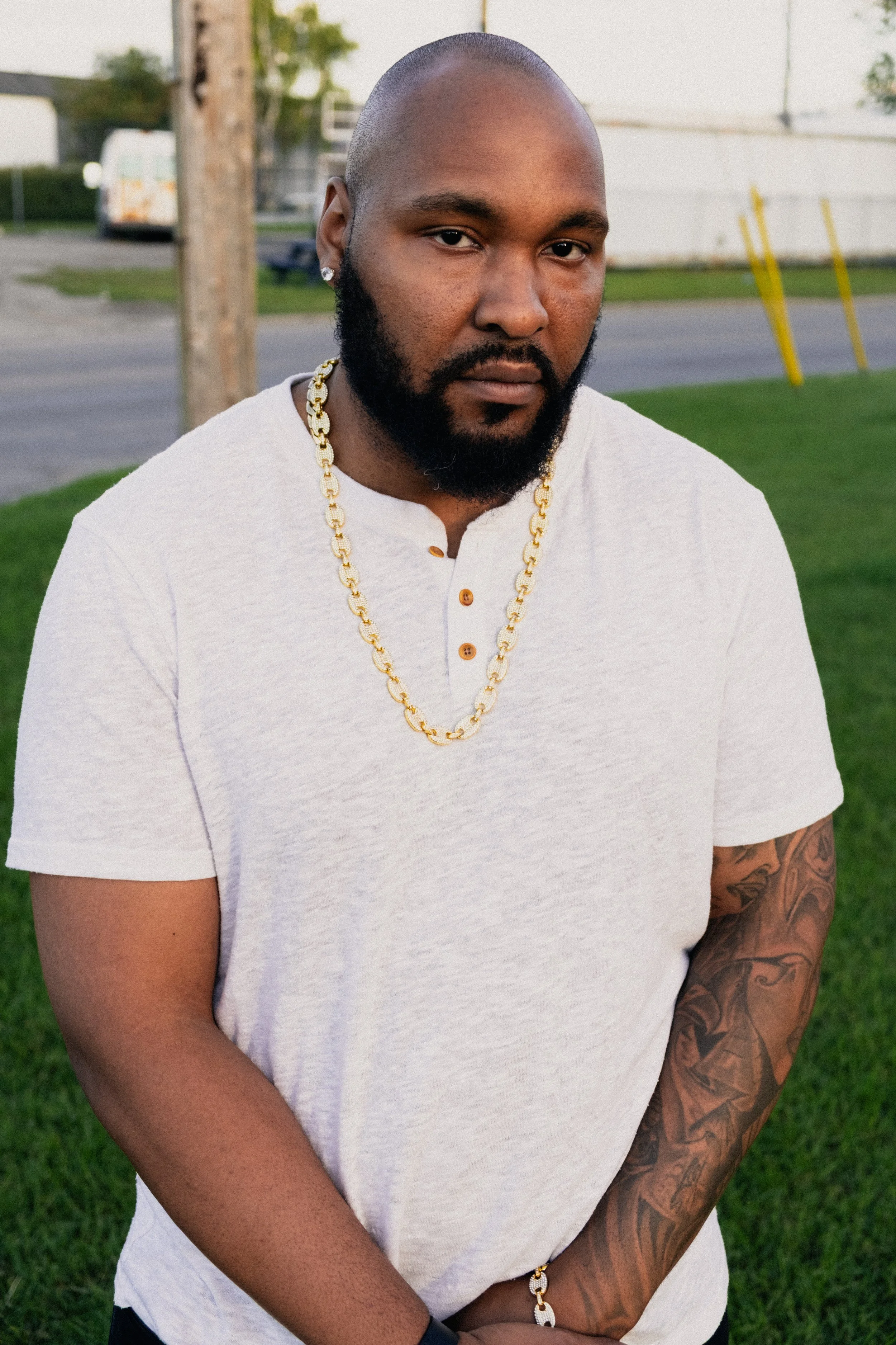 Portrait of a bearded Black man wearing a white shirt, gold chain, stud earring, and arm tattoos outdoors