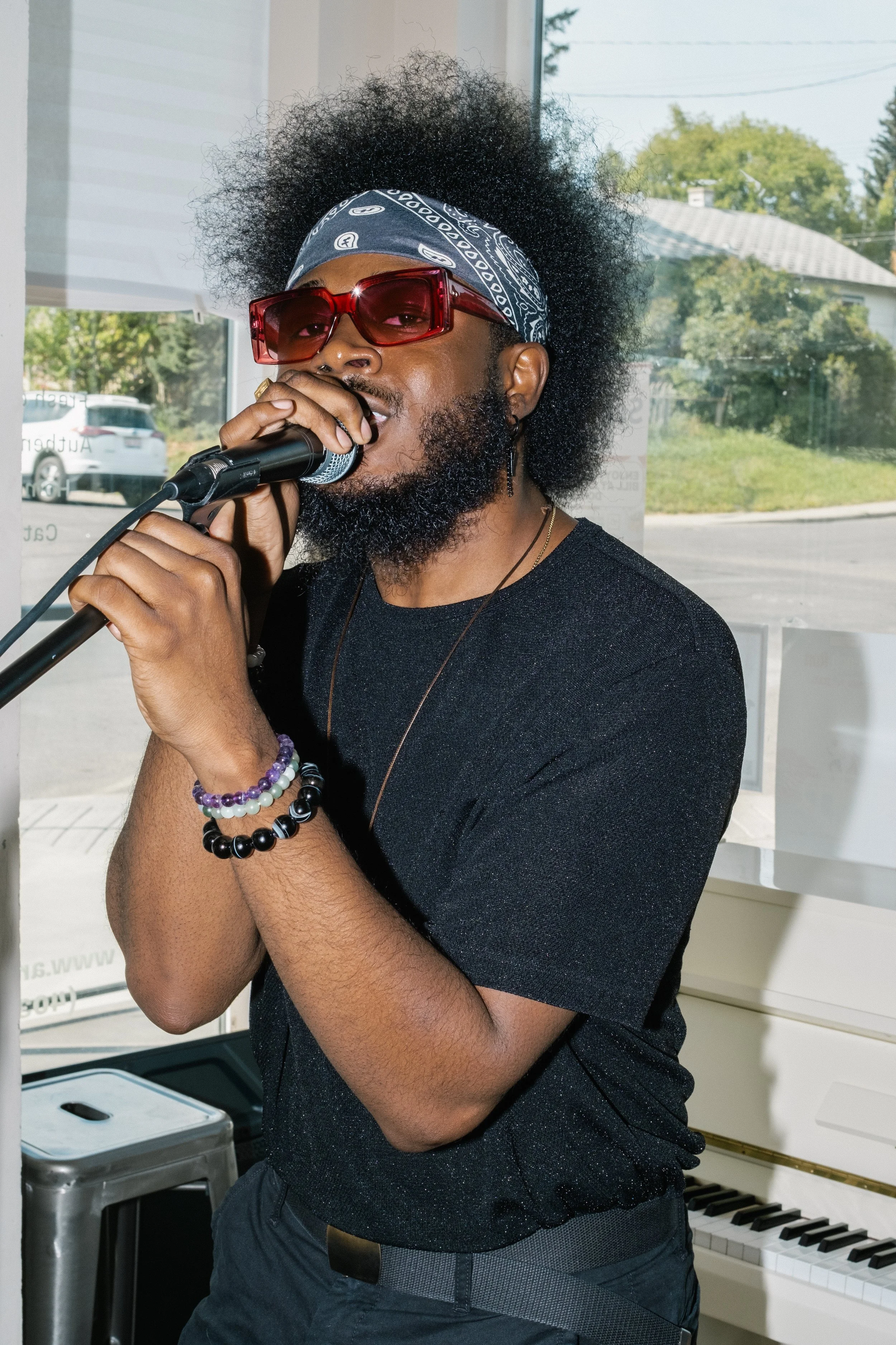 Teekay Othello wearing red sunglasses and a bandana, sings into a microphone in a room with a white piano and a large window showing trees and a street outside.