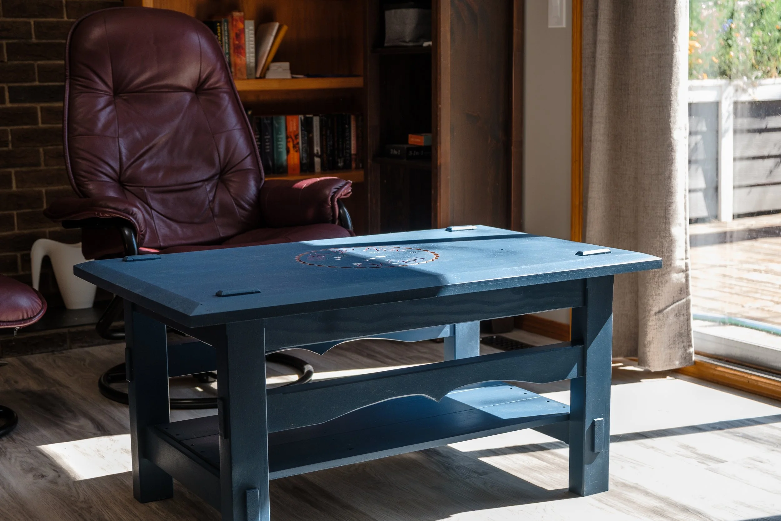 A blue table in a living room next to a burgundy leather office chair, with a bookshelf and window in the background.