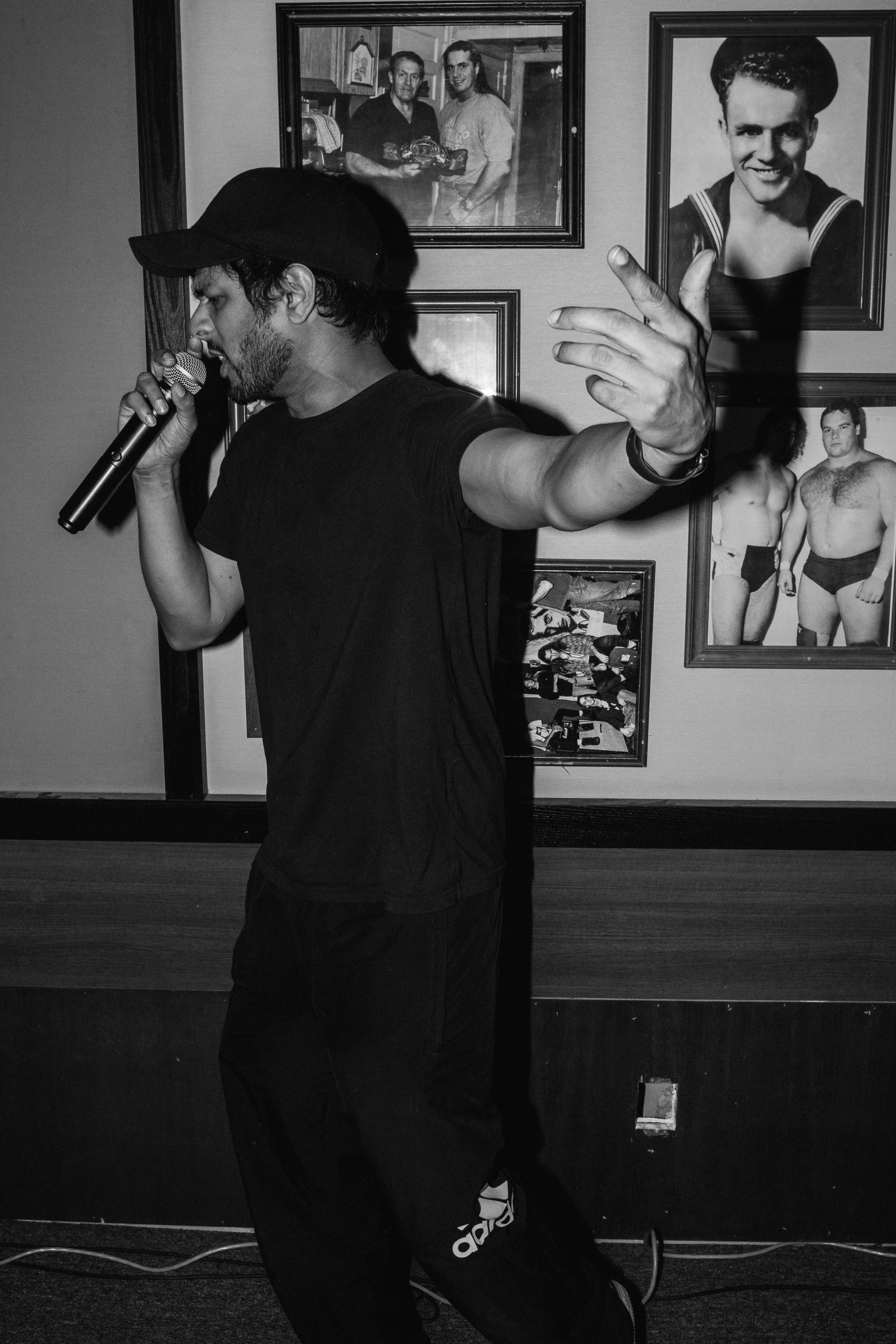HumLo frqntC singing into a microphone with wearing a baseball cap in a room with framed photographs of various people on the wall.