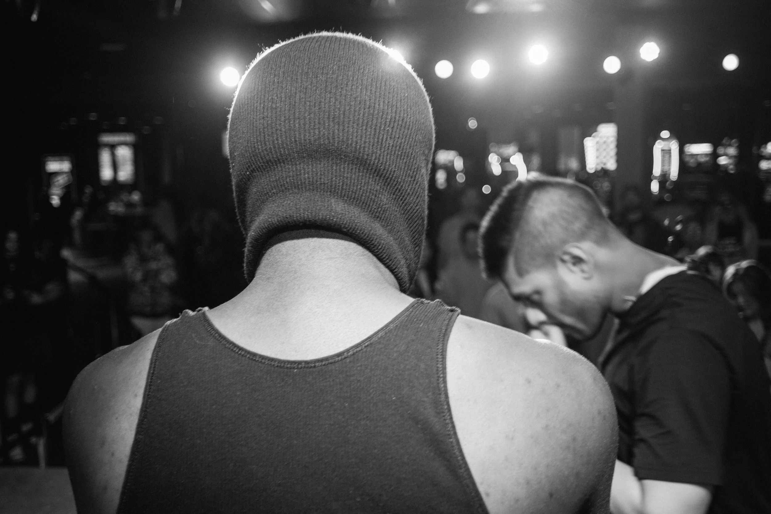 Black and white photo of a person with a beanie hat and sleeveless shirt, with their back to the camera, HumLo frqntC performing infront of them in a dimly lit indoor setting with bright lights overhead and other people in the background.