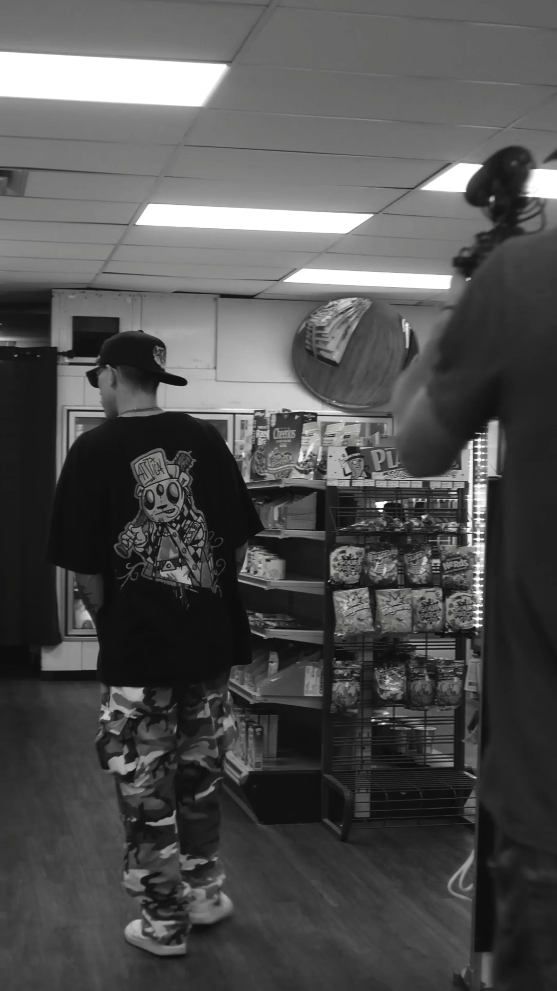 A black and white photo of two individuals in a store. One person is facing away, wearing a baseball cap, a graphic t-shirt, camouflage pants, and sunglasses. The other person, partially visible, is wearing a headset. Shelves with snacks and magazines are visible in the background.