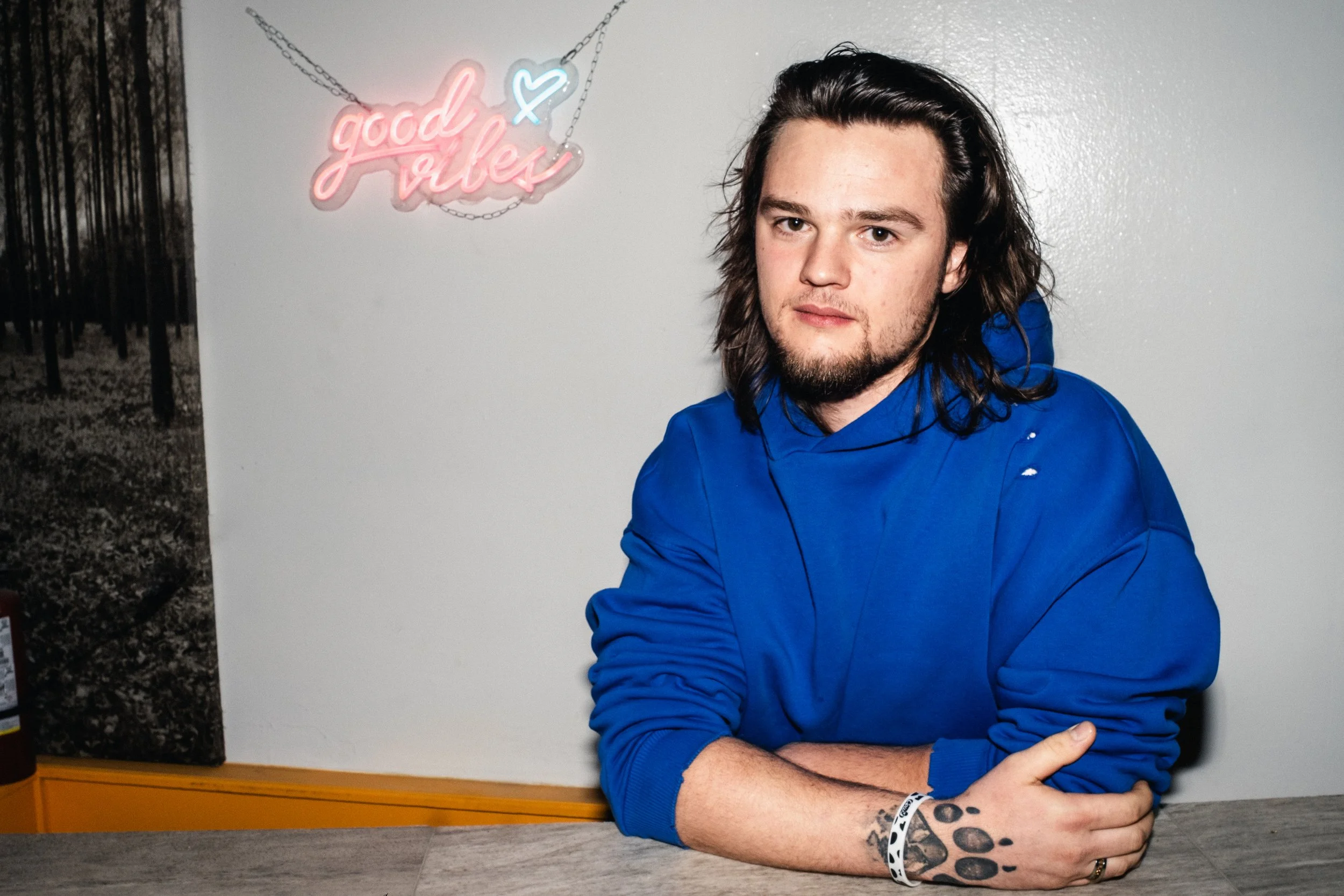 Artist Portrait: "Sexy Ryan" A man sitting with his arms crossed on a high table with long hair wearing a blue hoodie and a light up sign in the background saying "good vibes".