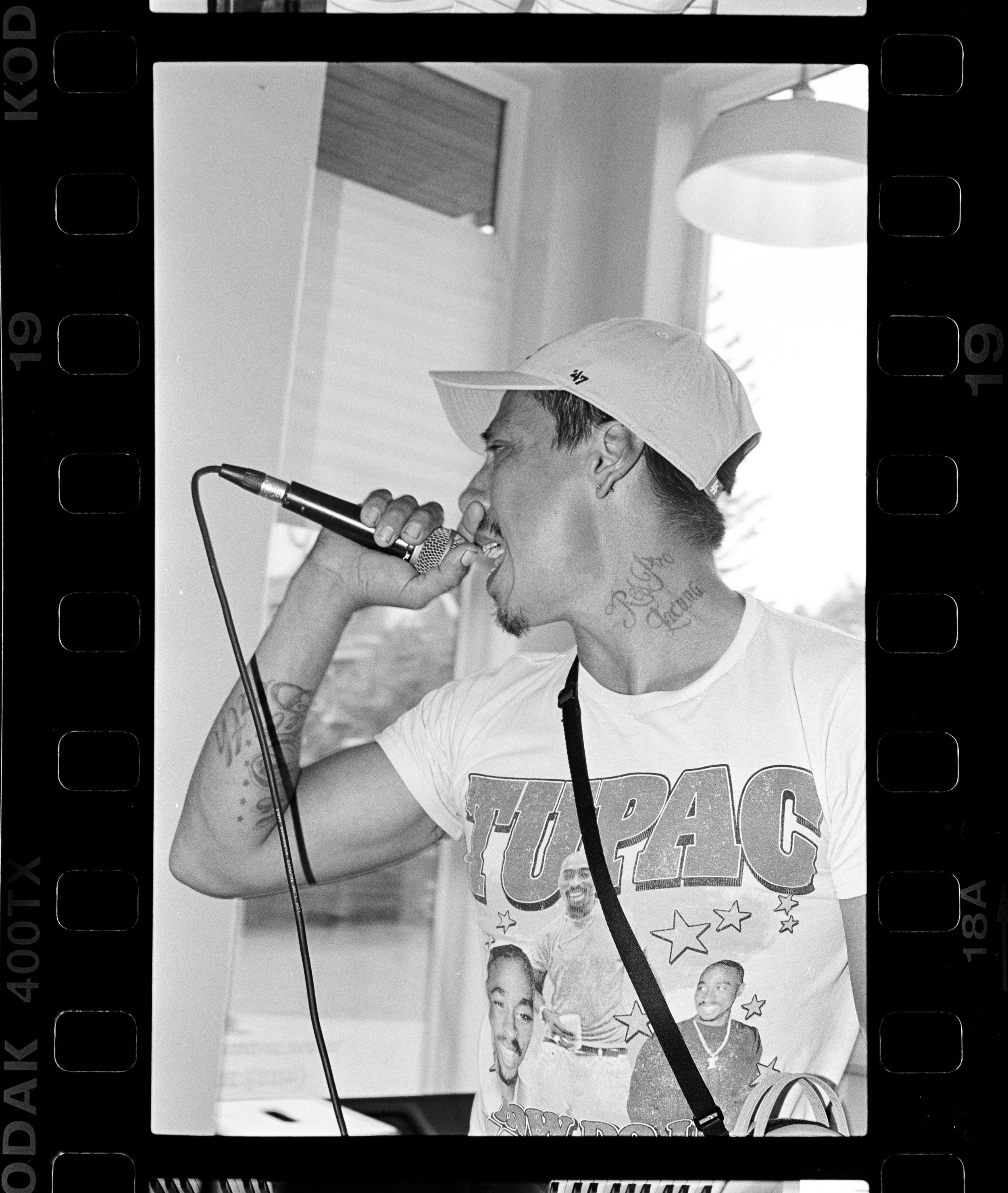 Tecun B'alam singing into a microphone, wearing a cap backwards and a Tupac shirt, with tattoos visible on his arm and neck, indoors near a window with blinds.