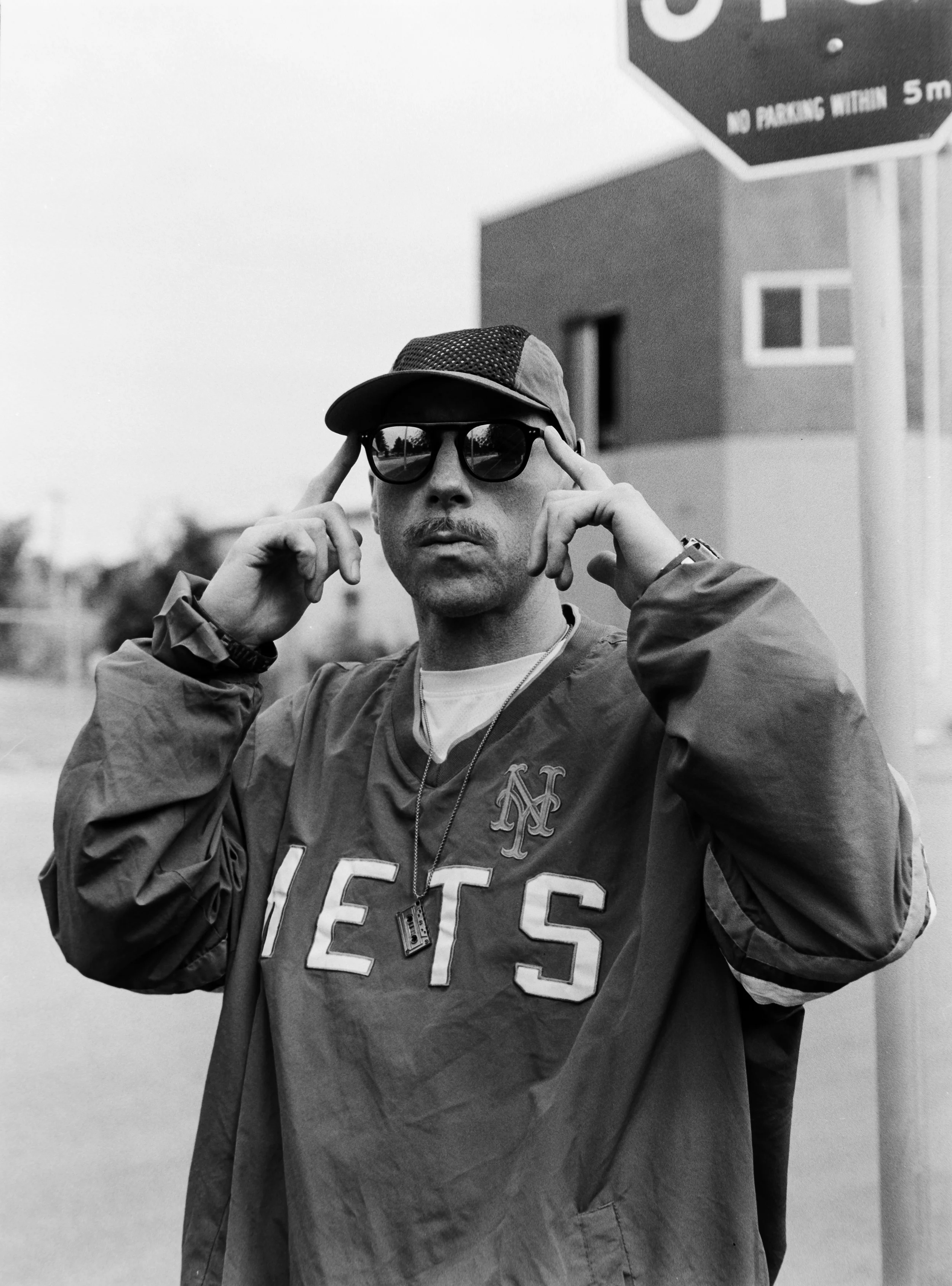 Artist Portrait: "THE ViNZ".  Wearing sports sunglasses, a cap, and a Mets jacket, standing outdoors near a stop sign, pointing to his sunglasses with both hands.