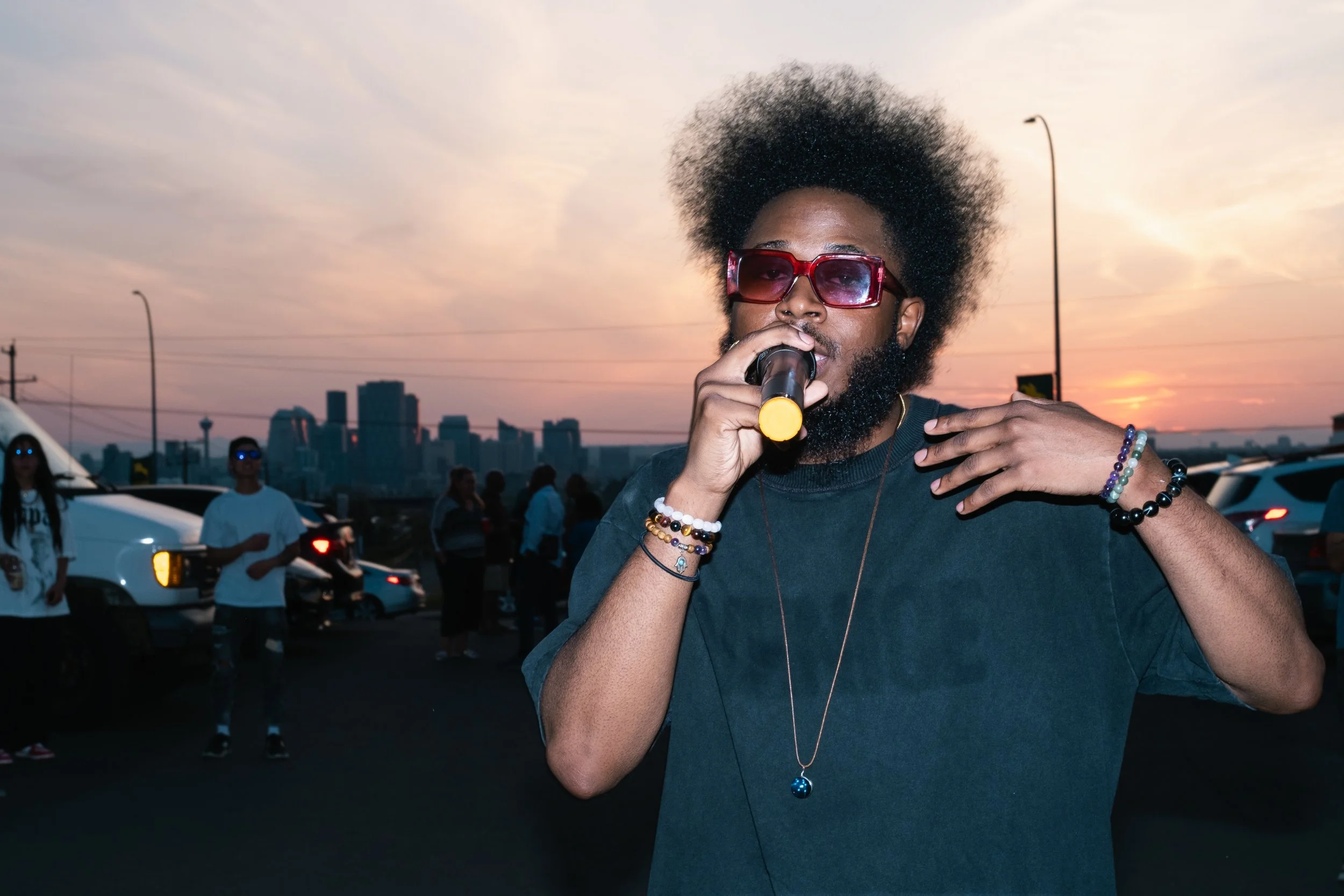 Teekay Othello with sunglasses and a beard holding a microphone, standing outdoors during sunset with a city skyline and parked cars in the background.