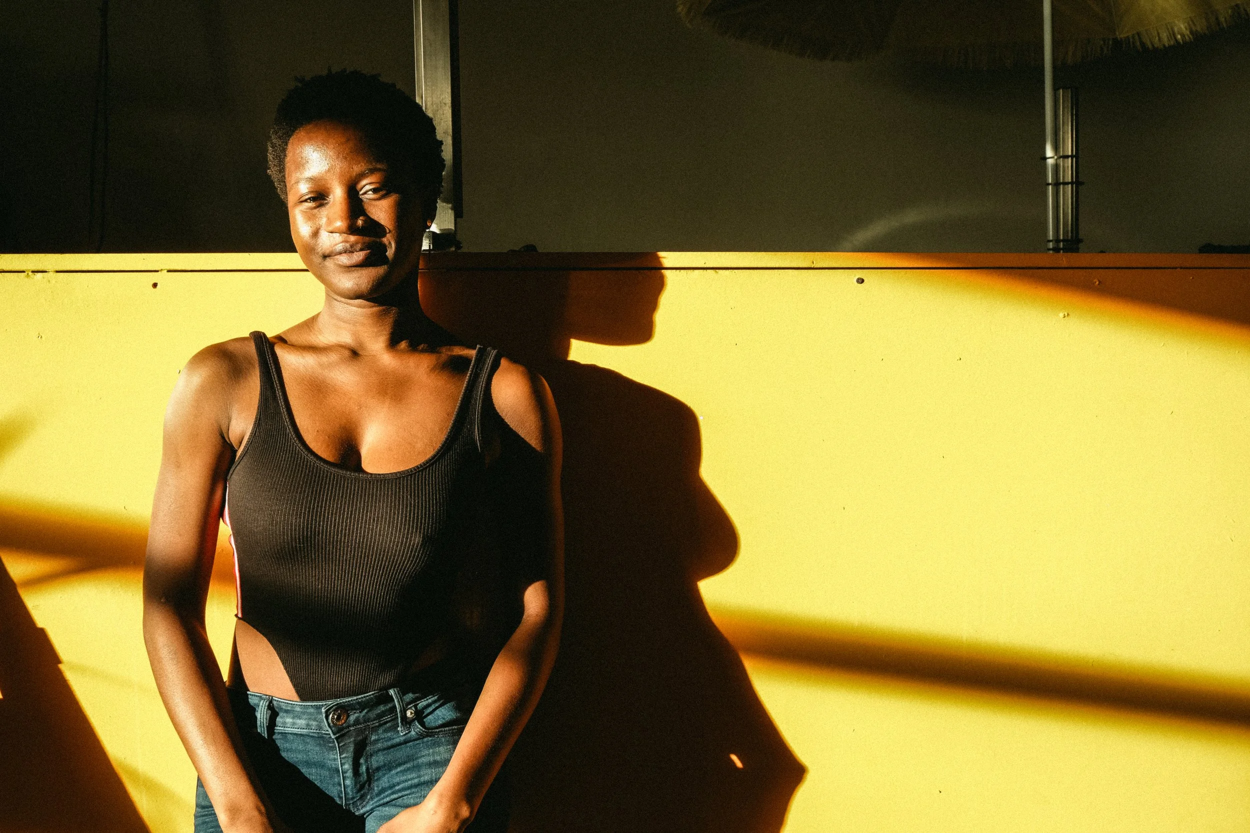 A woman with short curly hair wearing a black tank top and blue jeans, standing in front of a yellow wall with sunlight casting her shadow on the wall.