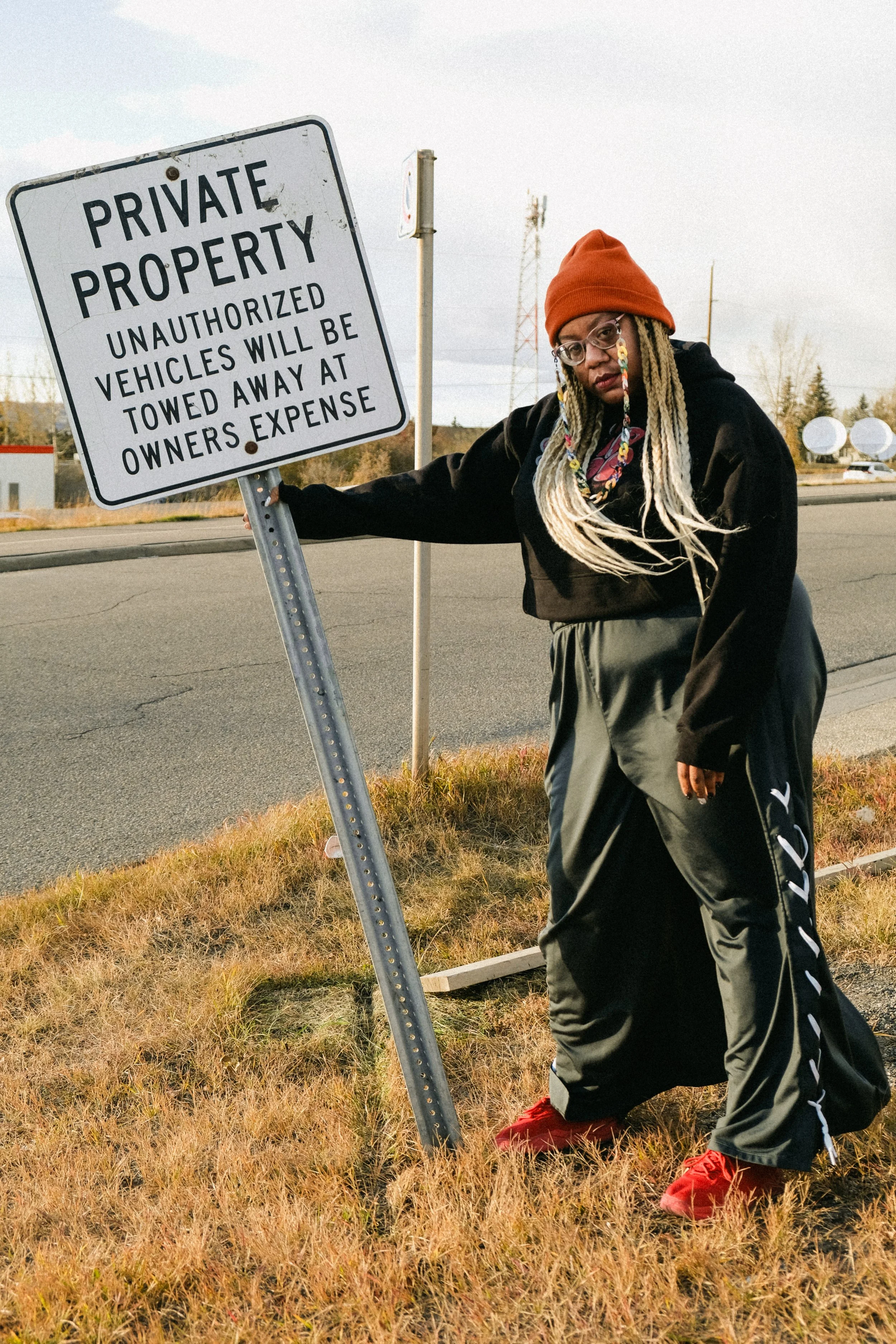 Artist Portrait: "Tea Fanny". Long blonde braids, red hat, glasses, black clothing, standing on grass beside a street, holding a "Private Property" sign that warns unauthorized vehicles will be towed at owner's expense.