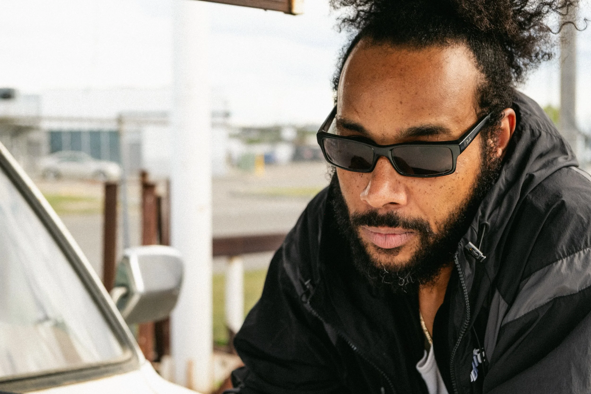 Artist Portrait: "Khanfidence". A man with curly hair and a beard wearing sunglasses and a black jacket outdoors near a vintage car.