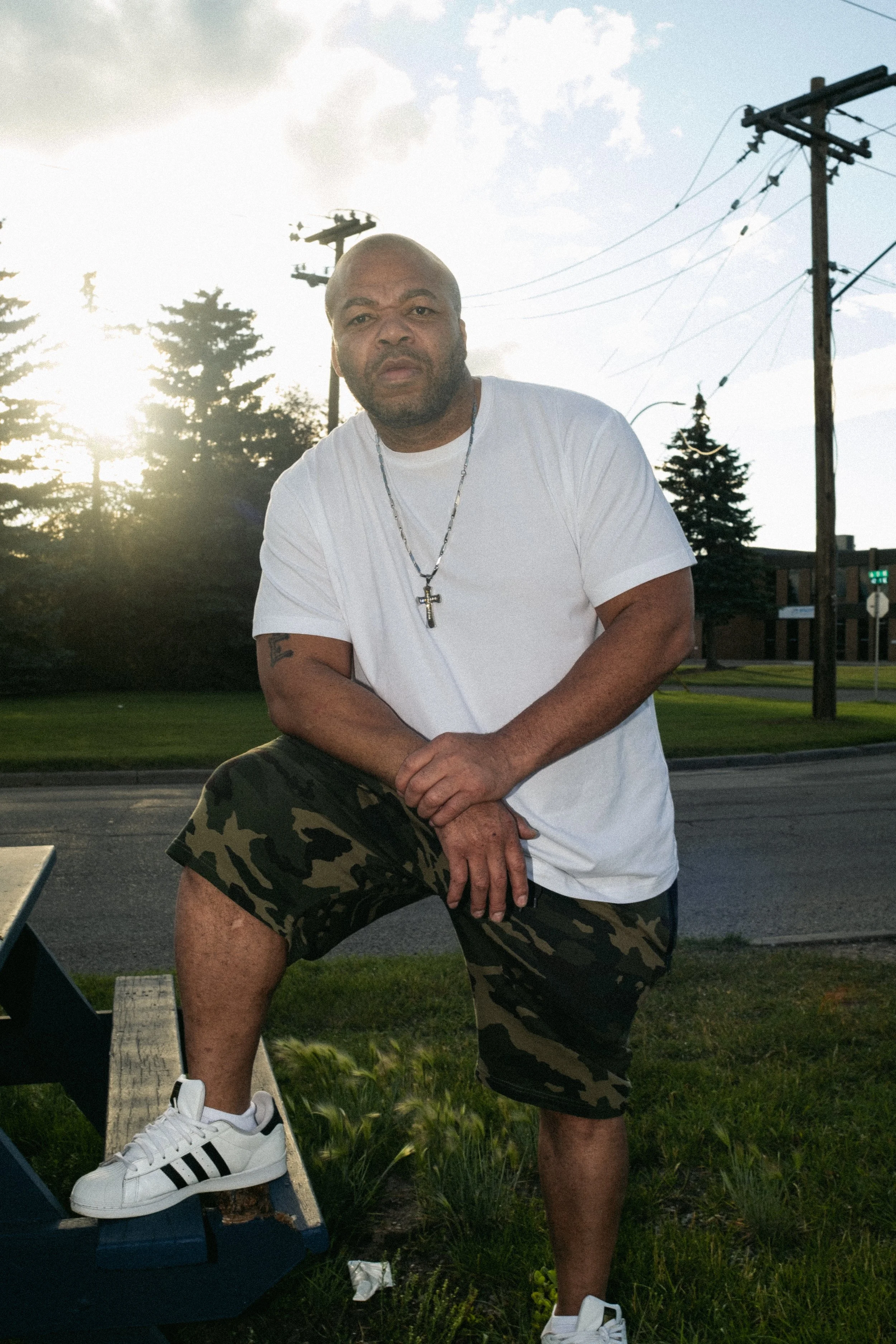 Artist Portrait: "Solomon Da Great". A man standing outdoors on a sidewalk with one foot on a bench, wearing a white T-shirt, camo shorts, and sneakers, with trees, power lines, and a partly cloudy sky in the background.