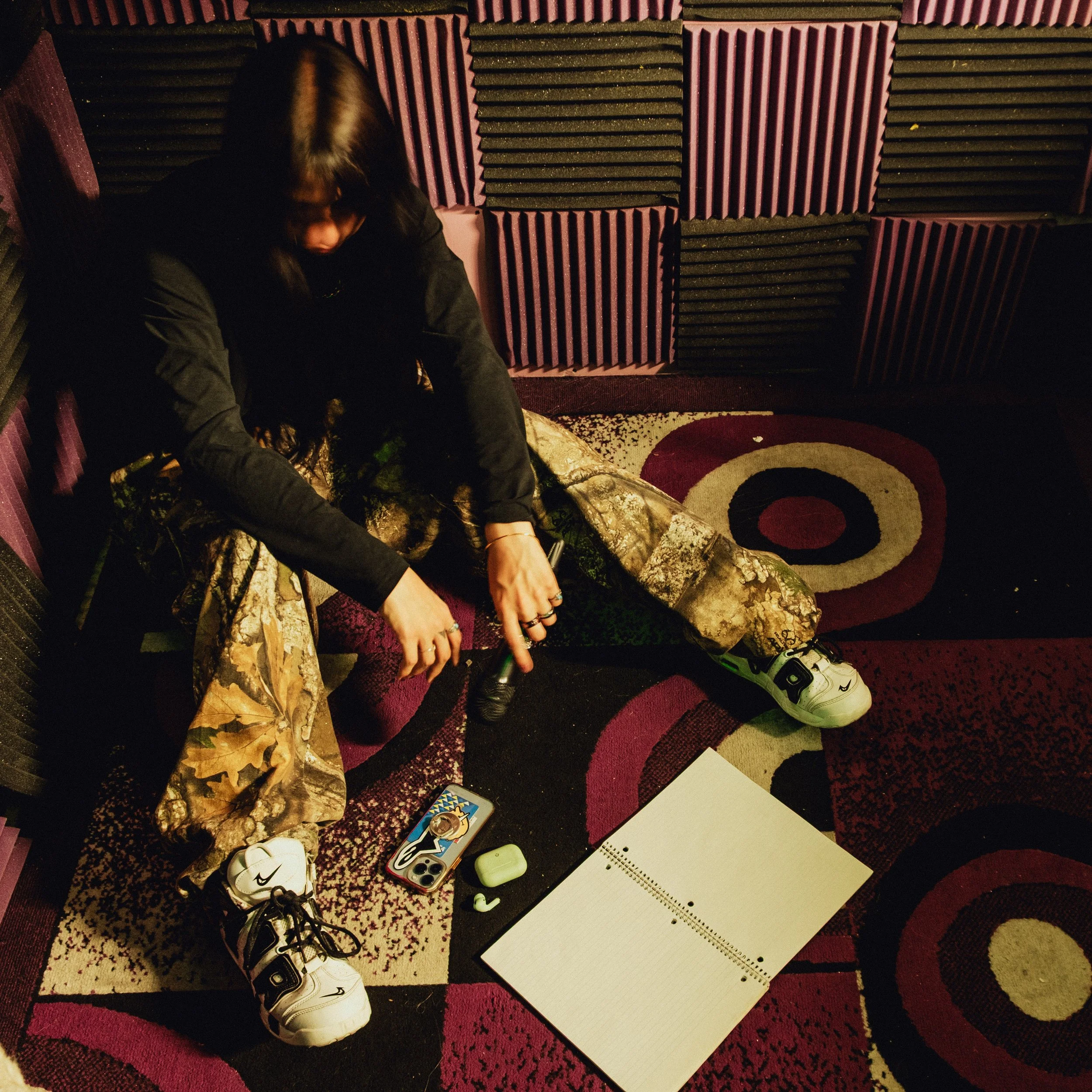 Rapper Lil $hawty / Lilmonawty Portrait sitting on the floor in a recording studio holding a microphone, with headphones, phone, a notebook, and earbuds nearby, surrounded by soundproof acoustic foam panels on the walls.