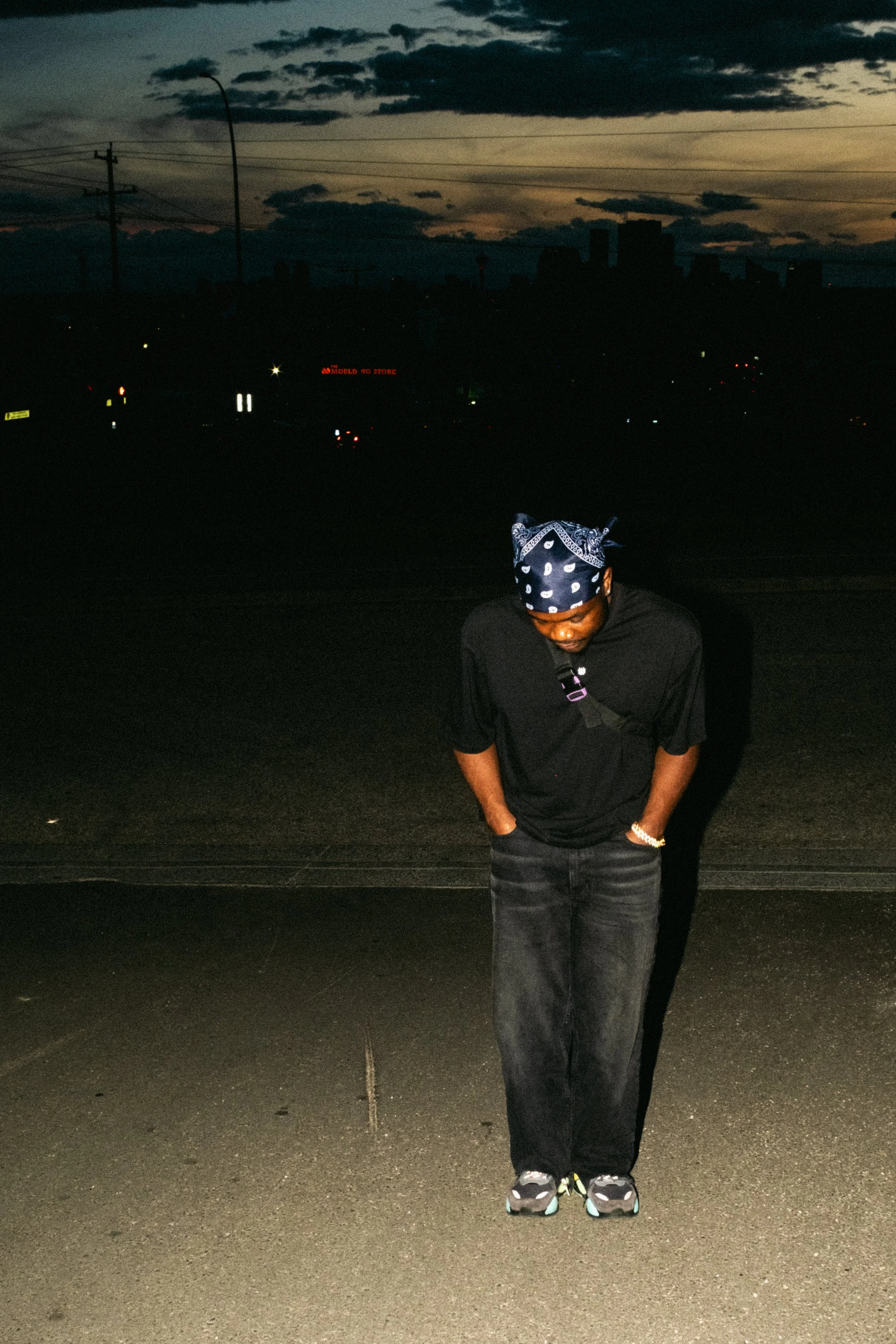 Artist Portrait: "Types". A person wearing a black t-shirt, dark jeans, a bandana, and sneakers standing on a dark street during dusk with a city skyline and some illuminated signs in the background.