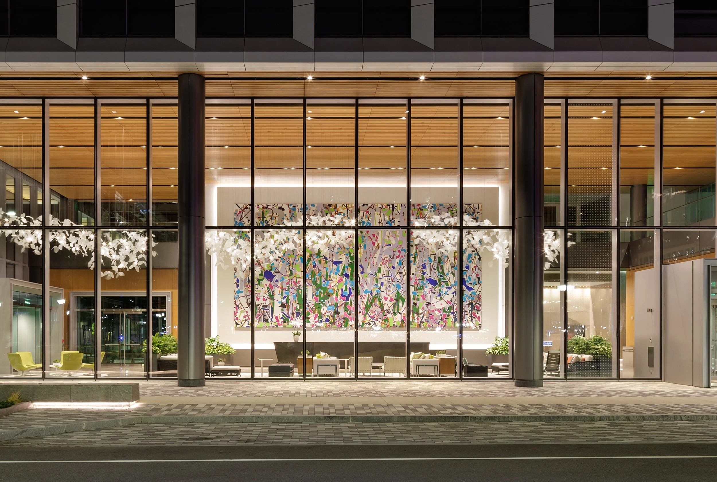 Night view of a modern building with large glass windows, showing an interior with an extra large painting by Amy Ellingson, filling the wall of the lobby, white hanging light fixtures, and seating areas.