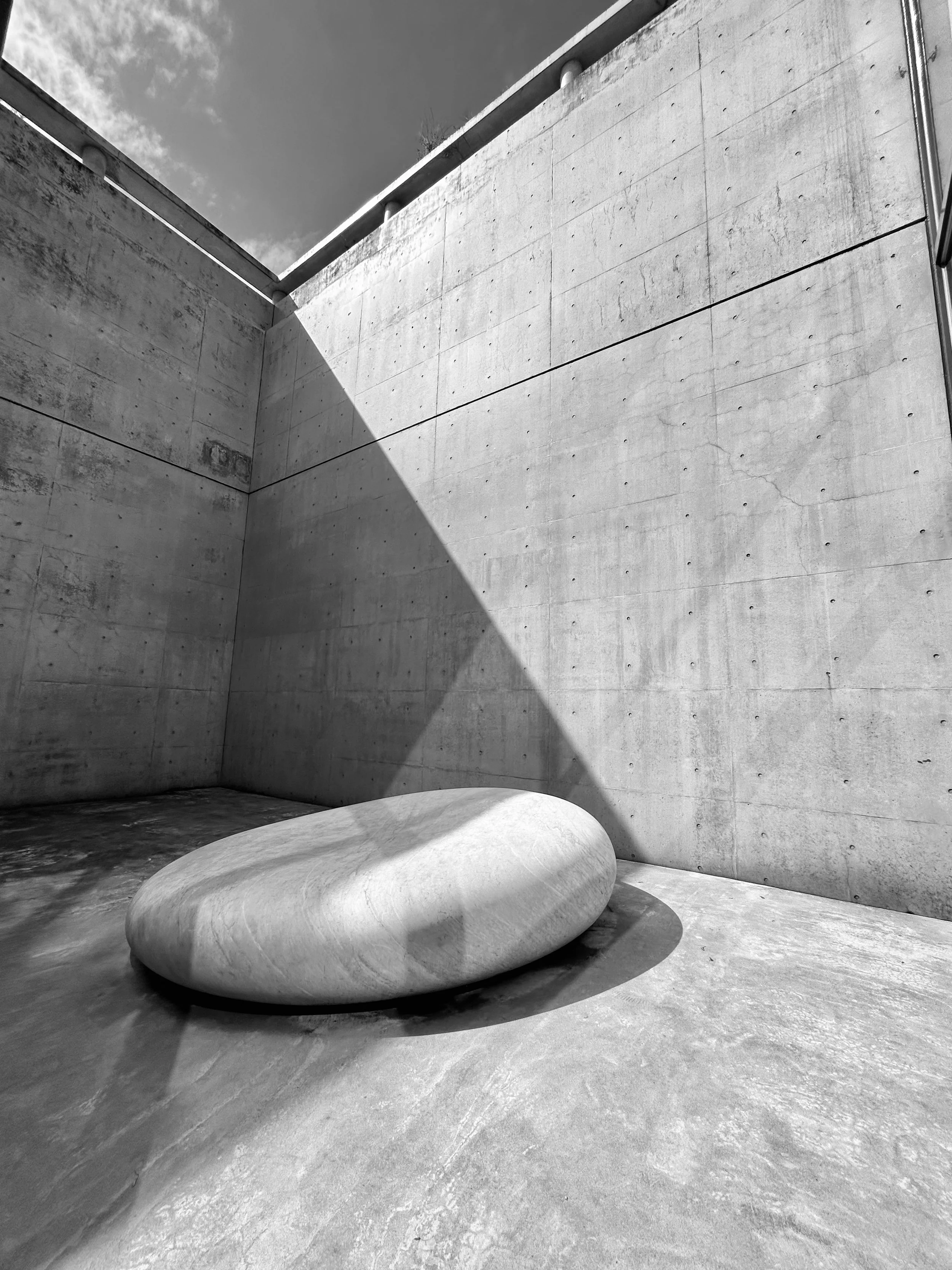 Minimalist outdoor space with concrete walls and floor, featuring a smooth, oval-shaped stone bench casting a shadow, under partly cloudy skies.