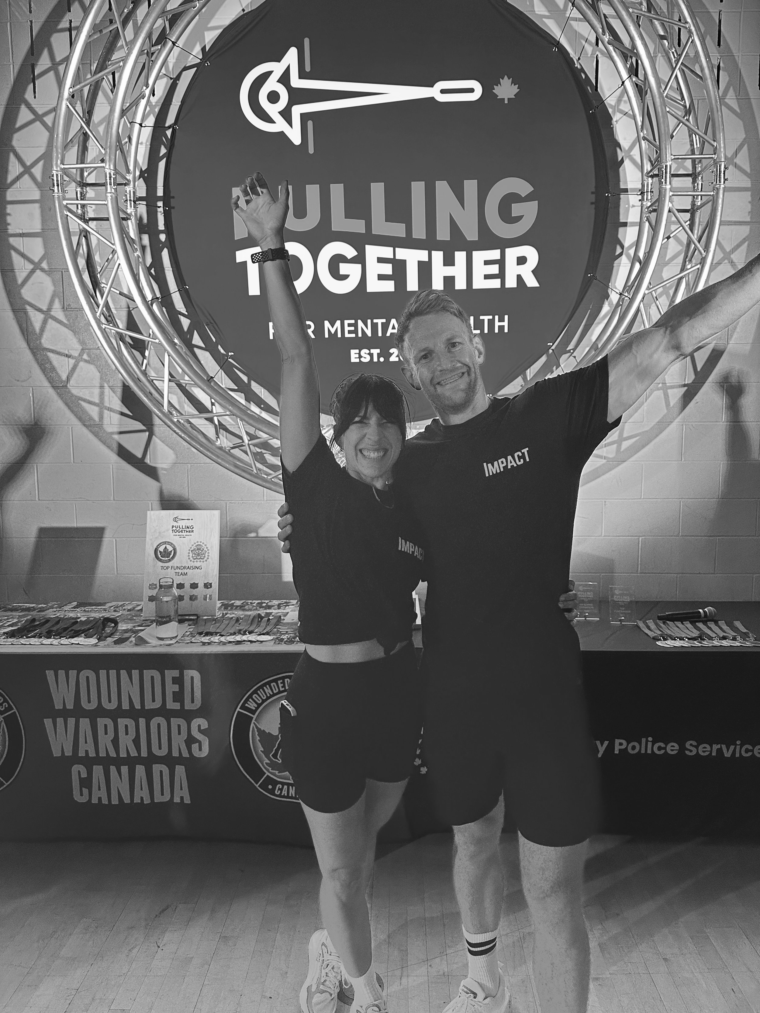 Two smiling people, a man and a woman, celebrating at a mental health awareness event called 'Pulling Together'. They are standing in front of a table with a banner that reads 'Wounded Warriors Canada'. The backdrop features a circular sign with an arrow and a maple leaf. The woman has short dark hair, and the man has light hair; both are dressed in black athletic clothing.