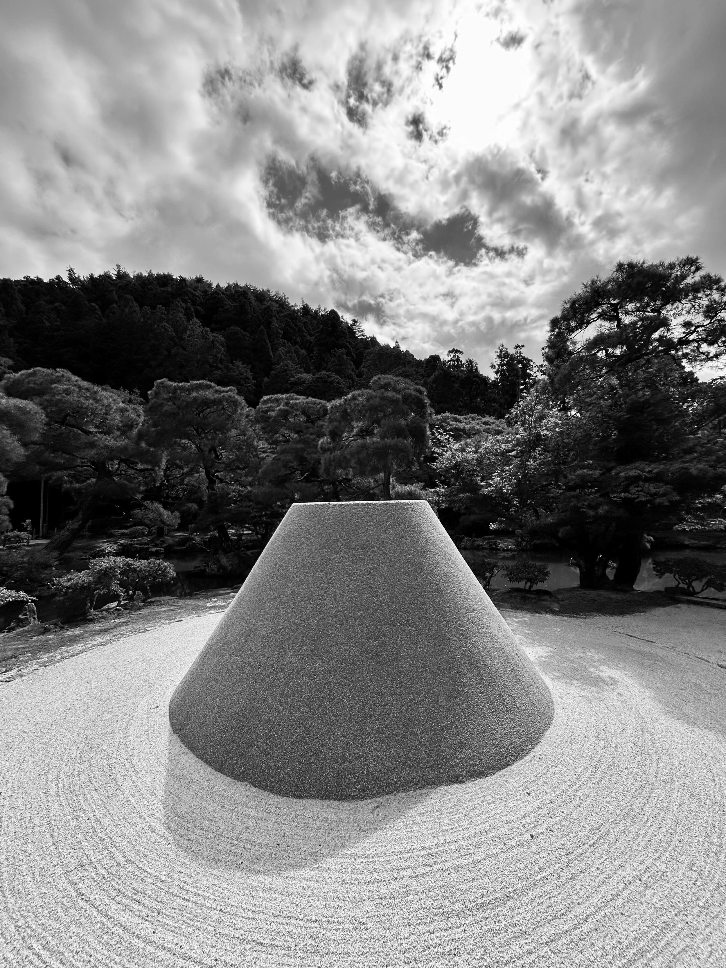 A large, cone-shaped stone sculpture in a Japanese Zen garden with neatly raked sand, surrounded by trees and hills under a cloudy sky.