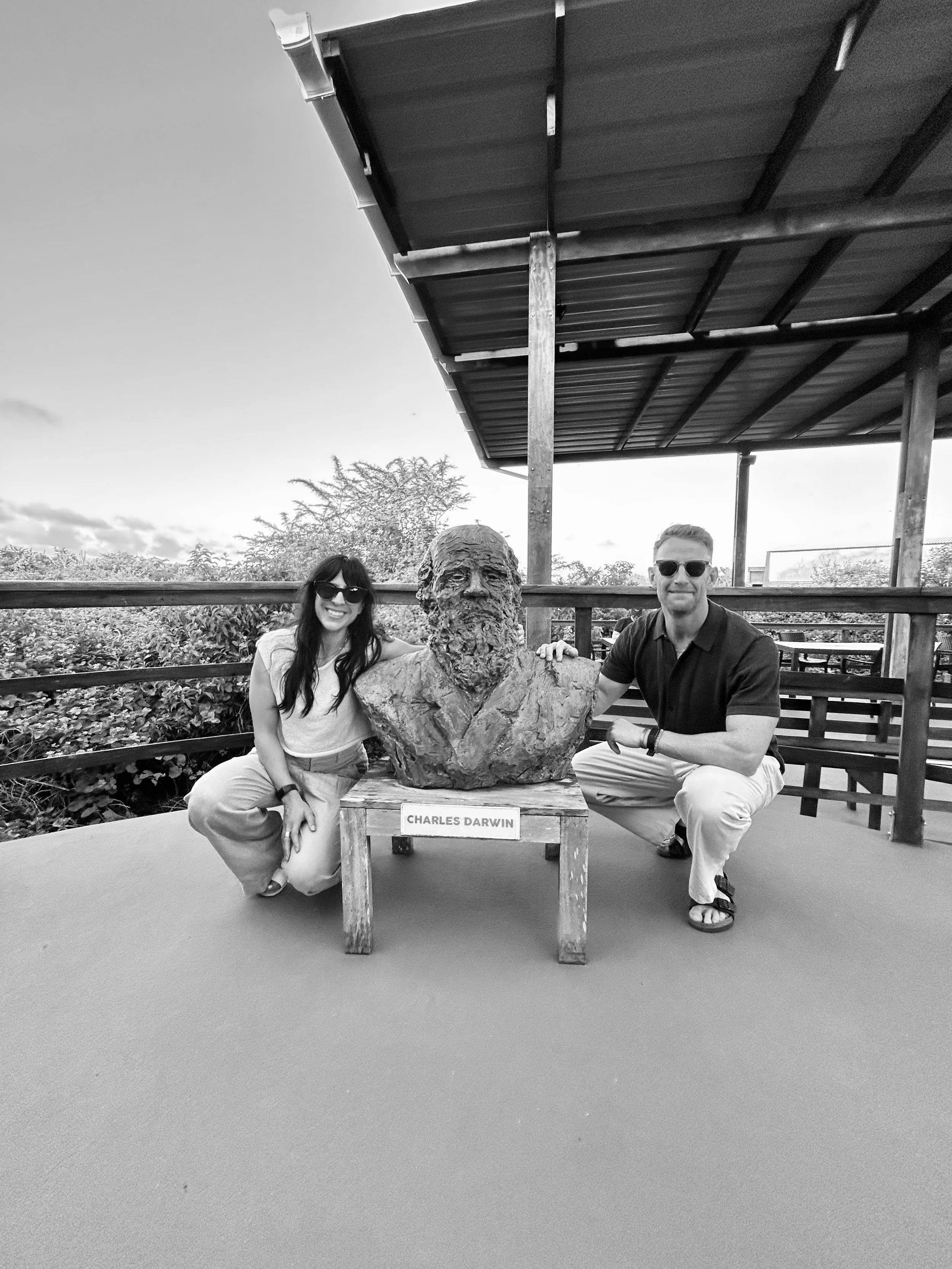 Jade and Rory kneeling next to a bust of Charles Darwin outdoors, with a sign reading 'Charles Darwin' on a bench in front of the bust.