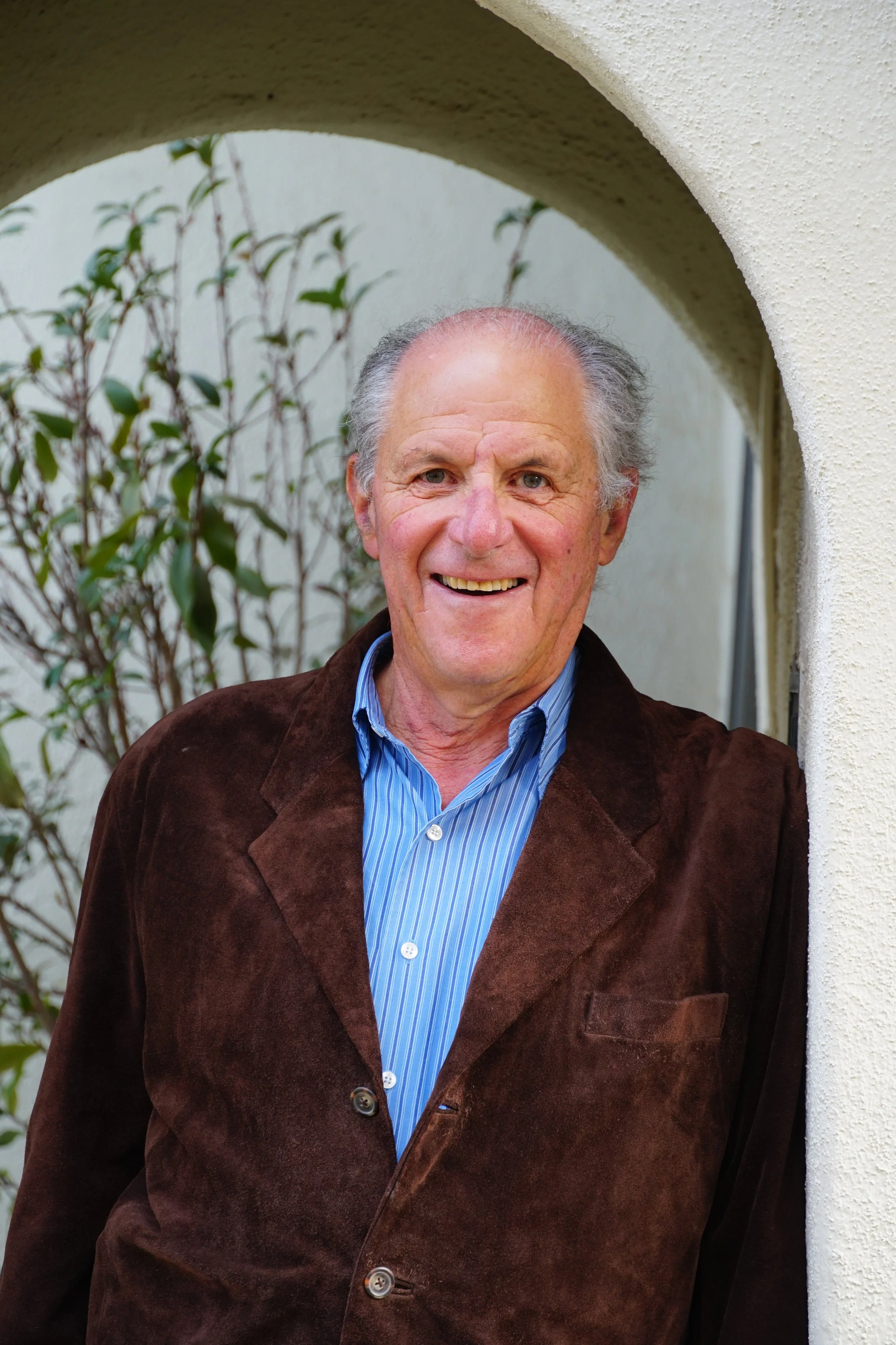 An elderly man with gray hair is smiling and standing in an archway, wearing a brown blazer and a blue striped shirt.