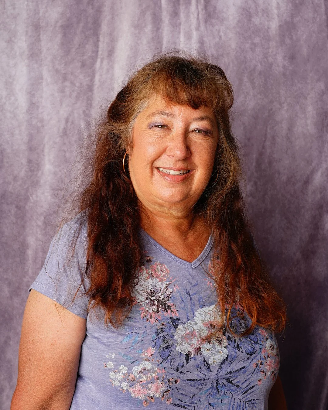 A young woman with shoulder-length black hair, smiling widely, wearing a dark blue top, outdoors with green foliage in the background.