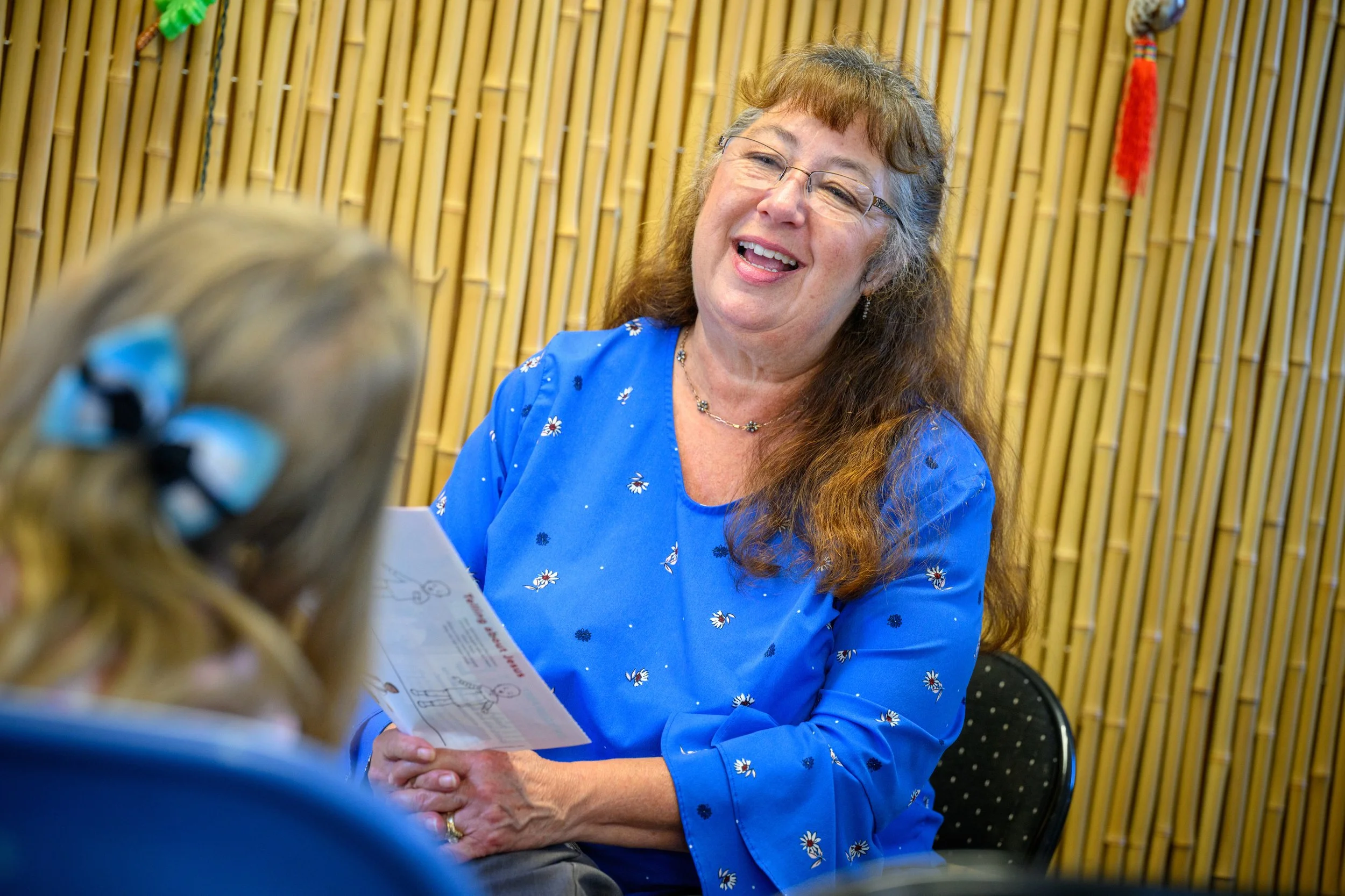 Woman with long wavy hair and glasses smiling while holding a paper, sitting in front of a bamboo wall, wearing a blue dress with small floral patterns.