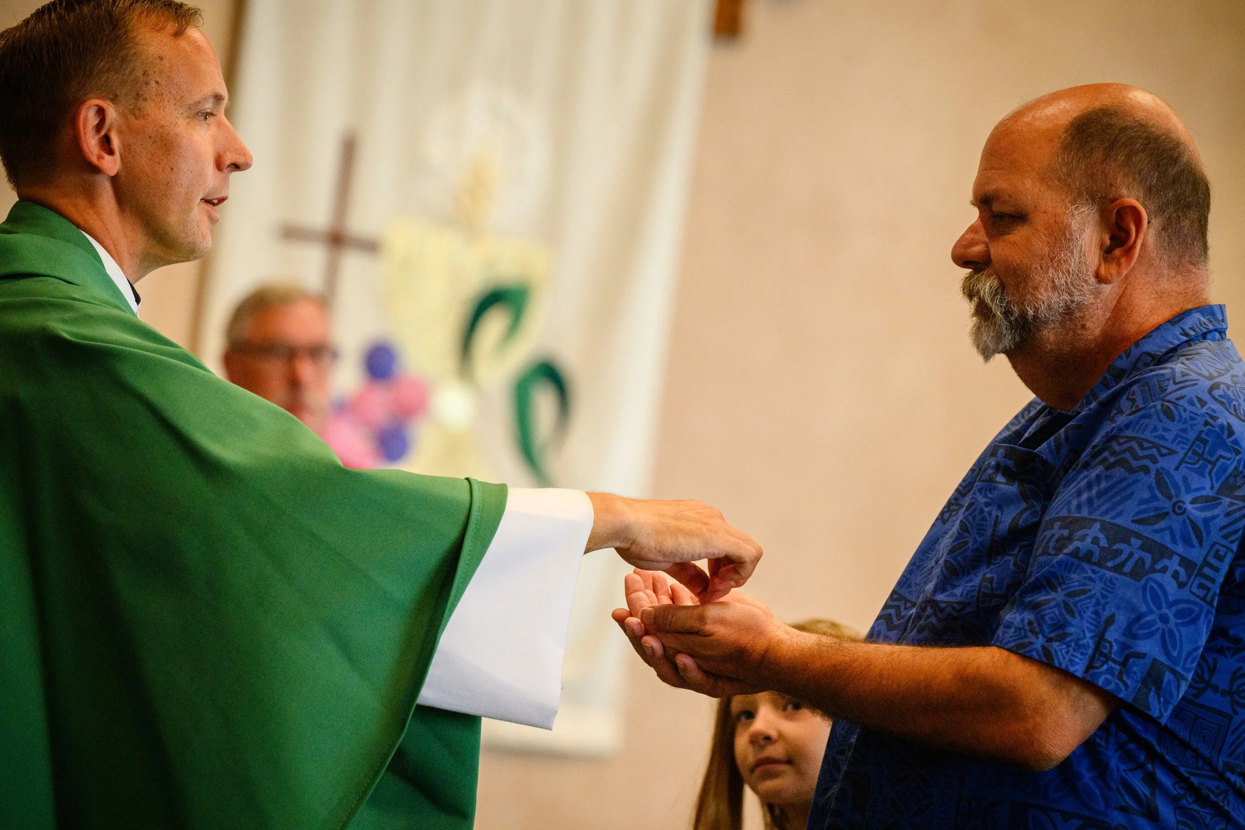 A priest dressed in green vestments is handing communion to a man with a beard wearing a blue Hawaiian shirt during a church service, with a young girl looking on in the background.