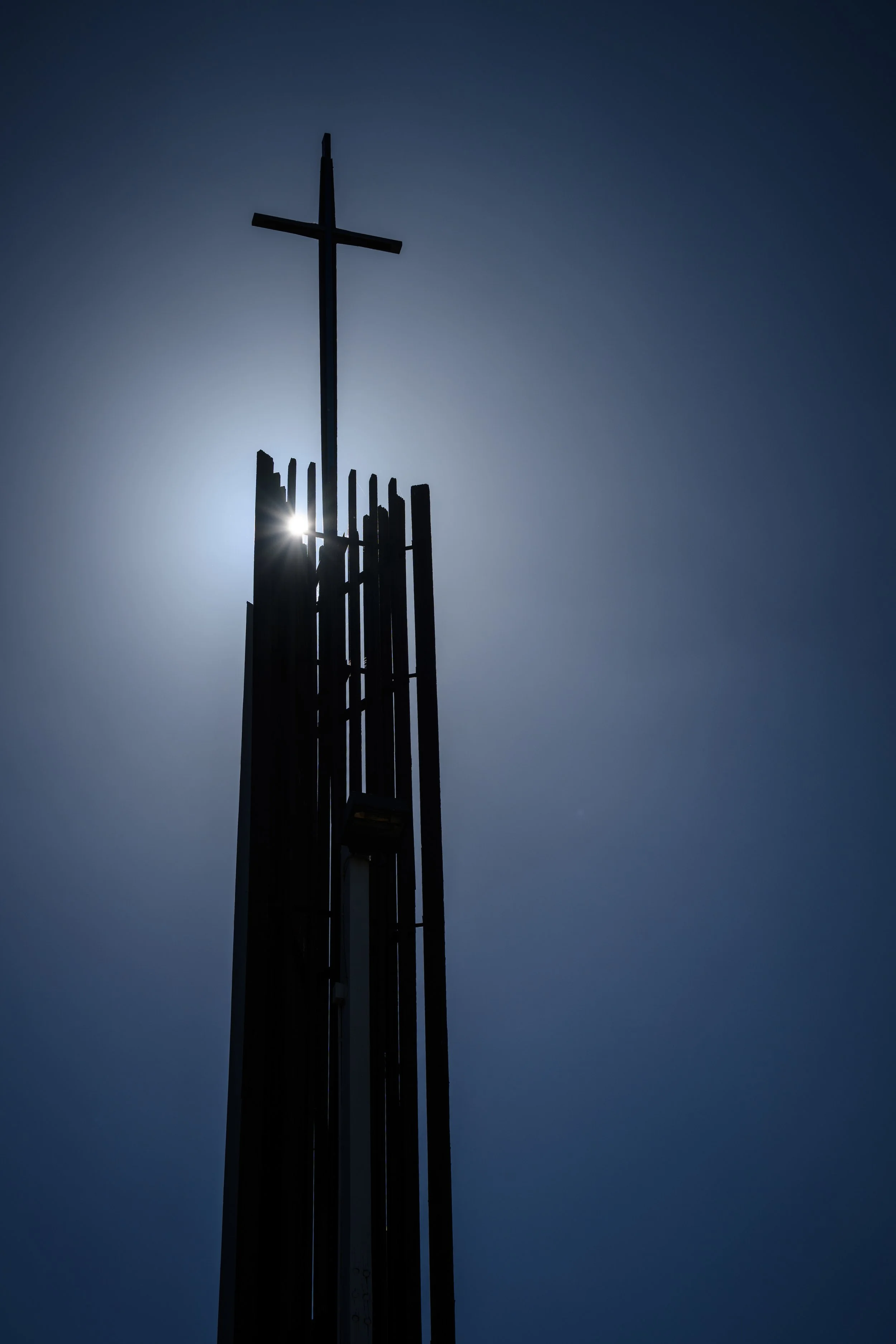A church steeple with a cross on top silhouetted against a bright sun and a clear blue sky.