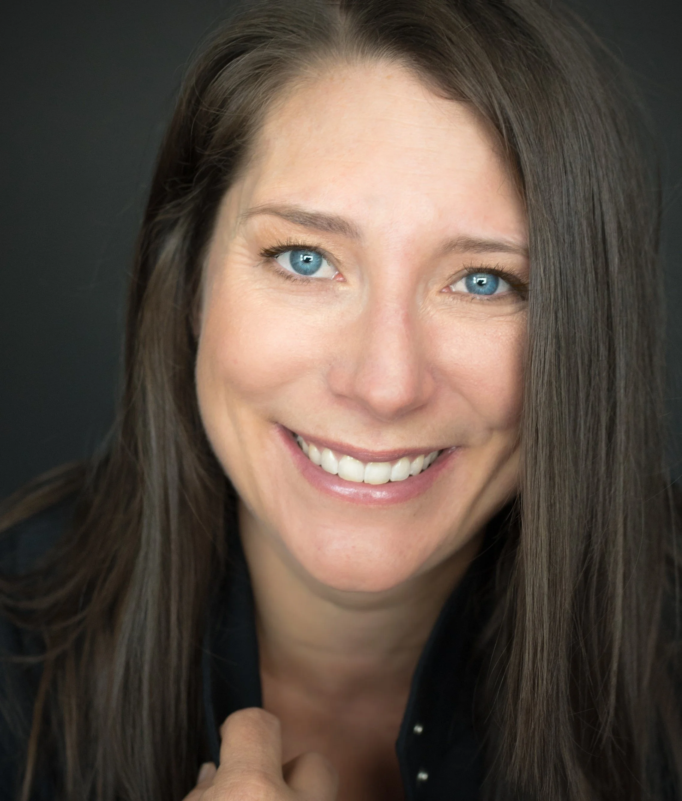 Close-up of a smiling woman with blue eyes and long brown hair, wearing a black top, against a dark background.