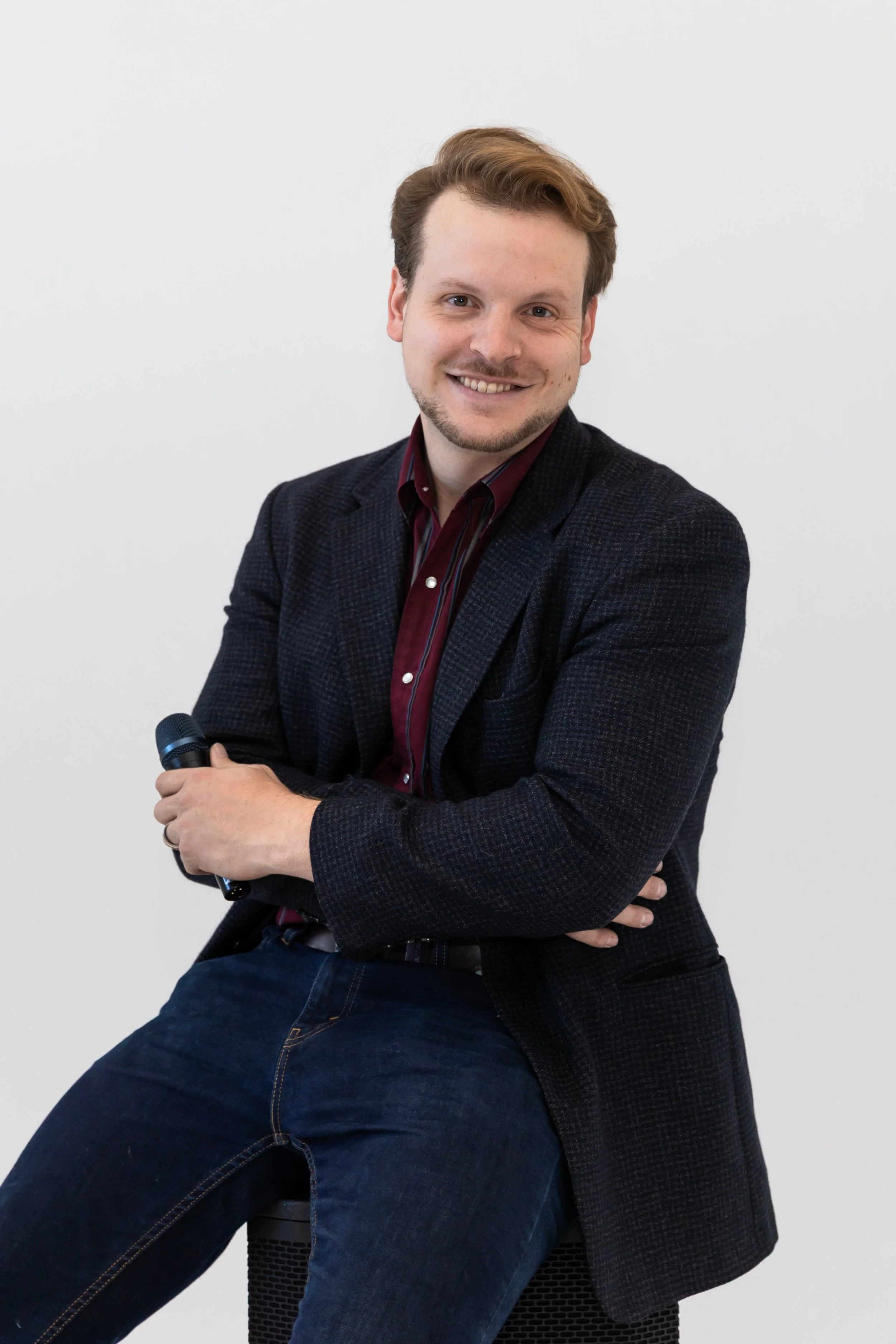 A smiling man with light skin, brown hair, and a beard, wearing a dark blazer over a red striped shirt, sitting on a stool, holding a microphone, against a plain white background.
