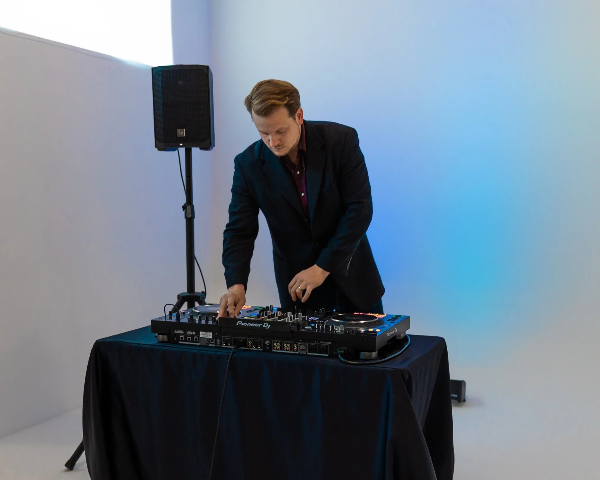 A man in a black blazer DJing with a Pioneer DJ controller on a black table, near a speaker, against a plain white wall with blue lighting.