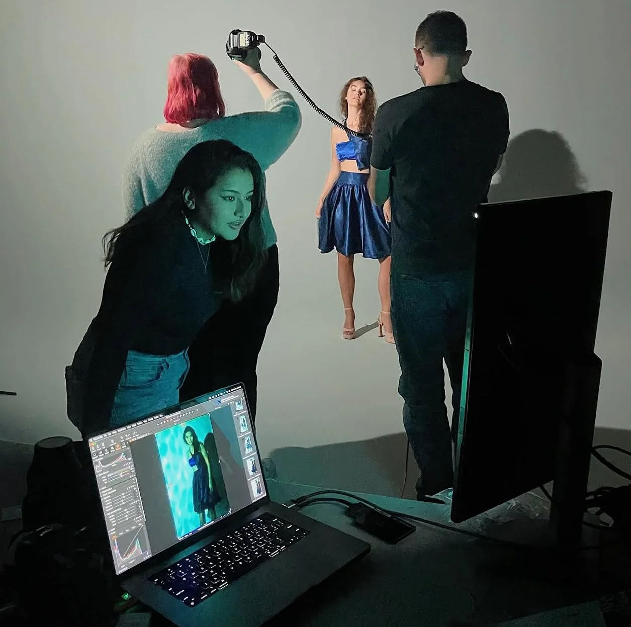 Behind the scenes of a photoshoot, a model in a blue outfit is being photographed by a photographer, with a videographer recording. A woman is reviewing images on a laptop, and a large monitor displays the model's picture. The setting suggests a professional studio environment.