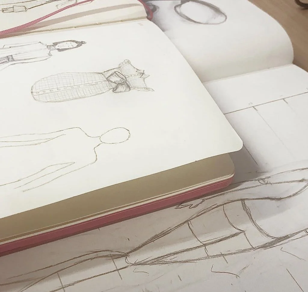 Open sketchbook with various fashion sketches, including a mannequin, a dressed figure, and a person sketch, placed on a wooden surface.