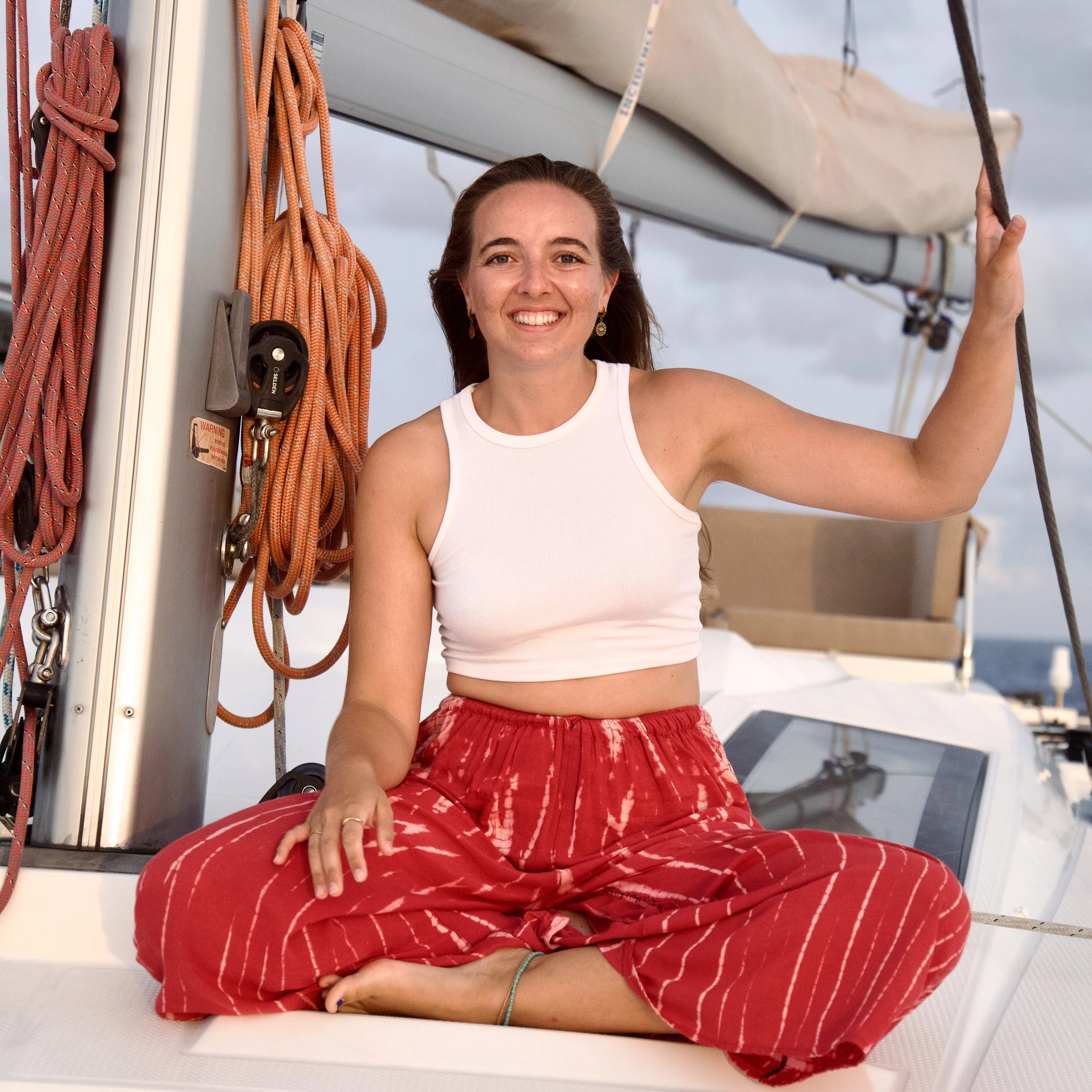 A woman with long brown hair, wearing a white sleeveless top and red tie-dye pants, sitting on a boat or yacht, smiling, holding her right hand up, and surrounded by sailing equipment such as ropes and a sail, with a cloudy sky in the background.