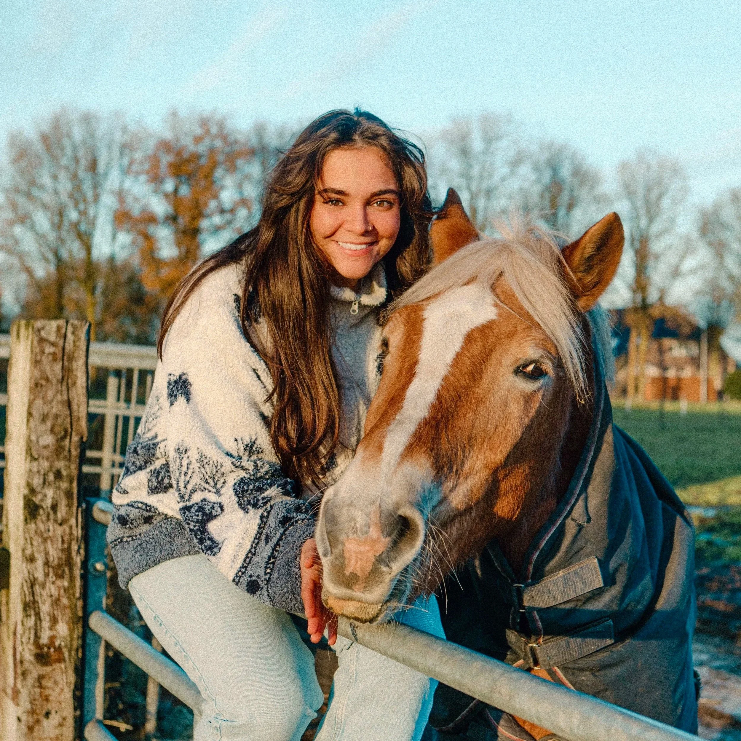 A young woman with long brown hair smiling while leaning on a chestnut horse with a white stripe on its face at an outdoor stable. The background includes leafless trees and a wooden fence.