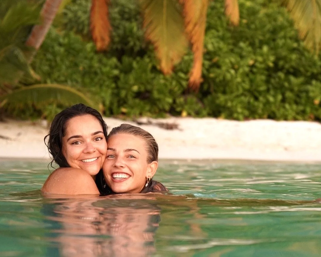 Two women smiling and hugging in the ocean near a sandy beach with palm trees and lush greenery in the background.