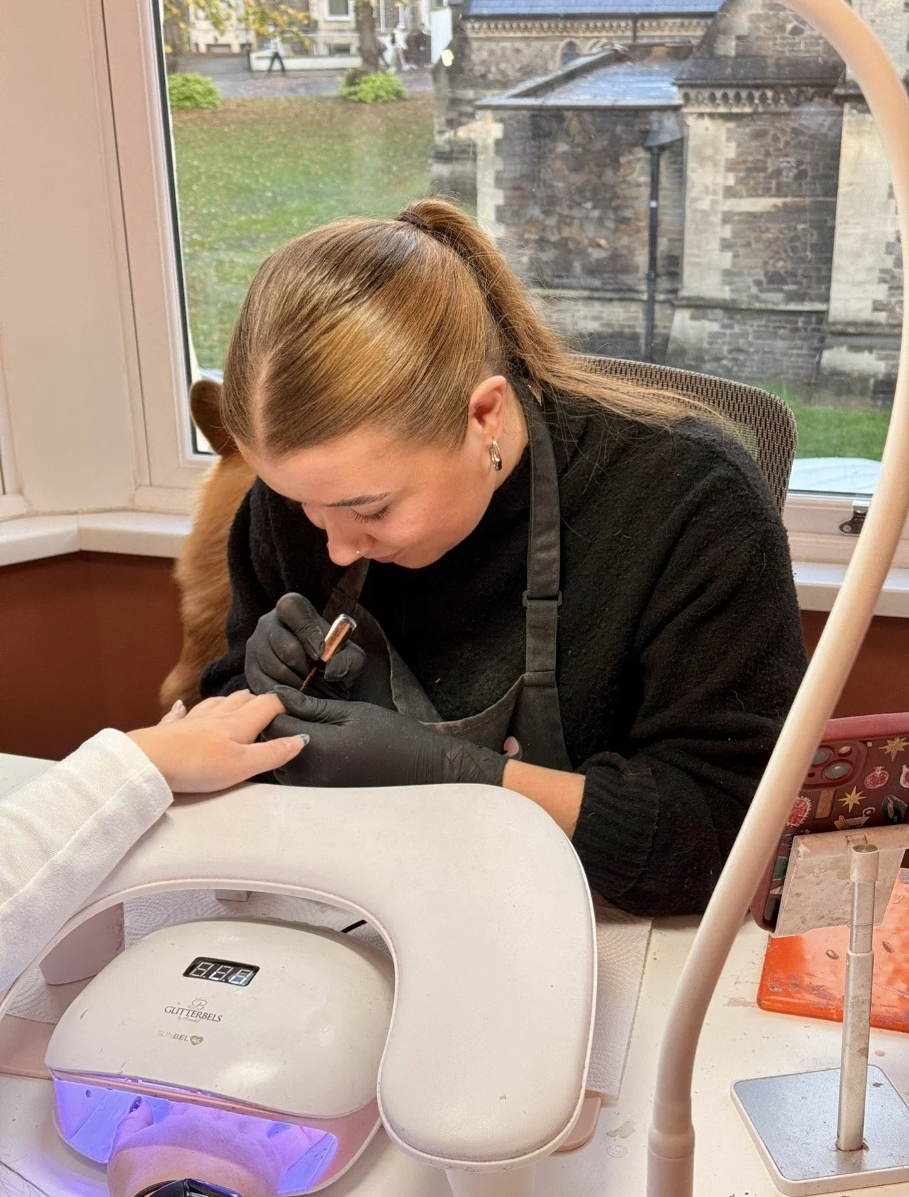 A woman getting a manicure at a nail salon, with a UV light curing the nails and a customer’s hand resting on the table.