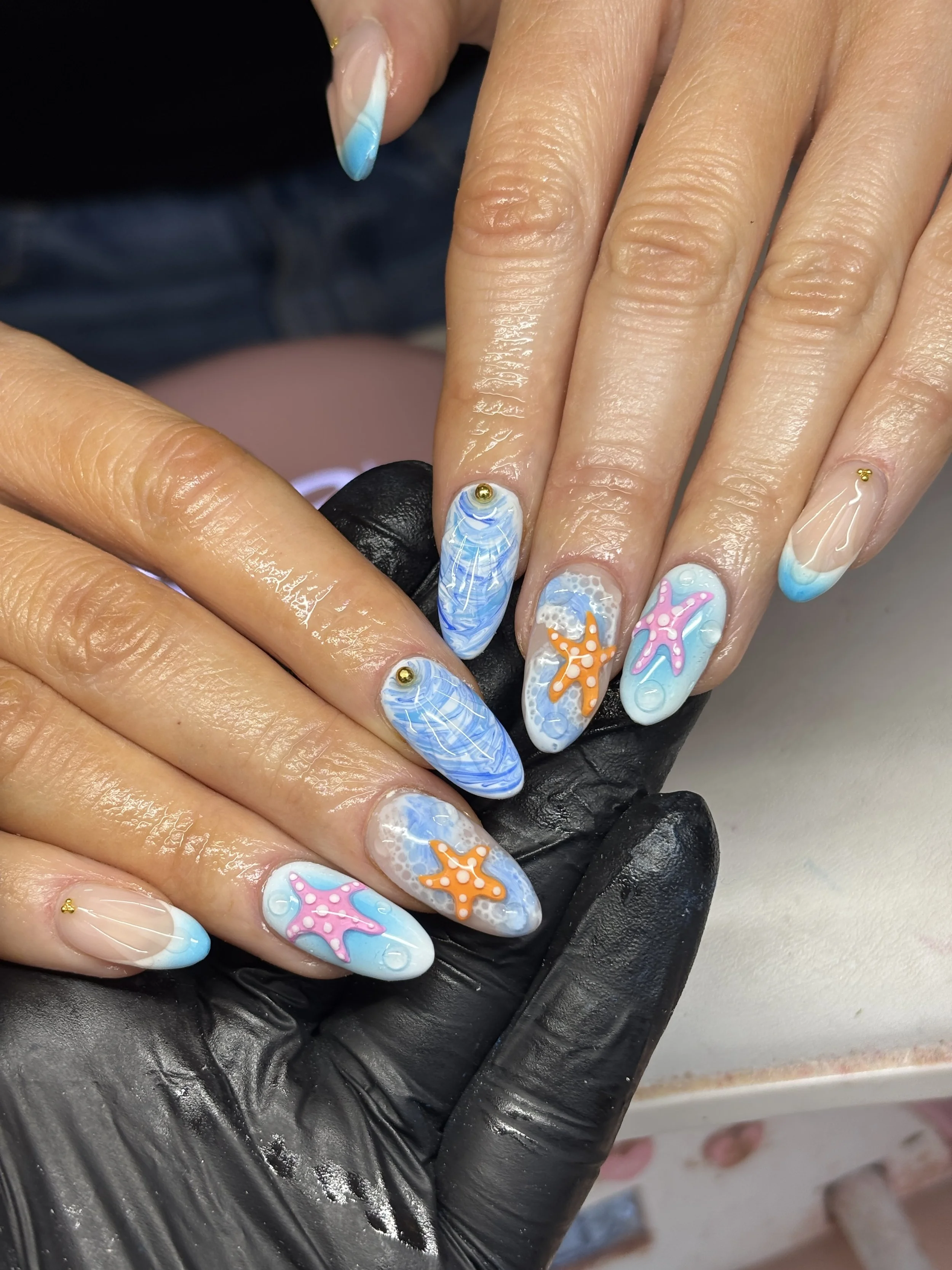 Nail art with a beach and ocean theme, featuring starfish and seashell designs in blue, pink, and orange, with gold accents.