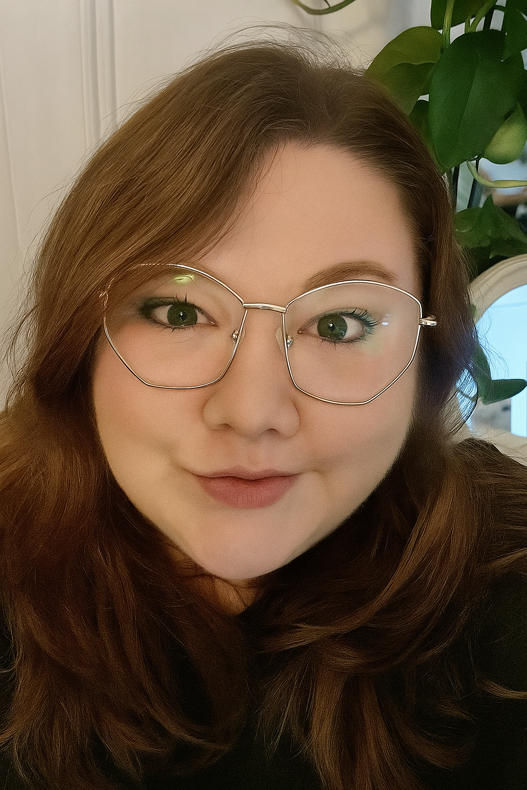 A woman with long, wavy brown hair wearing large, geometric glasses, posing for a selfie indoors with a houseplant in the background.