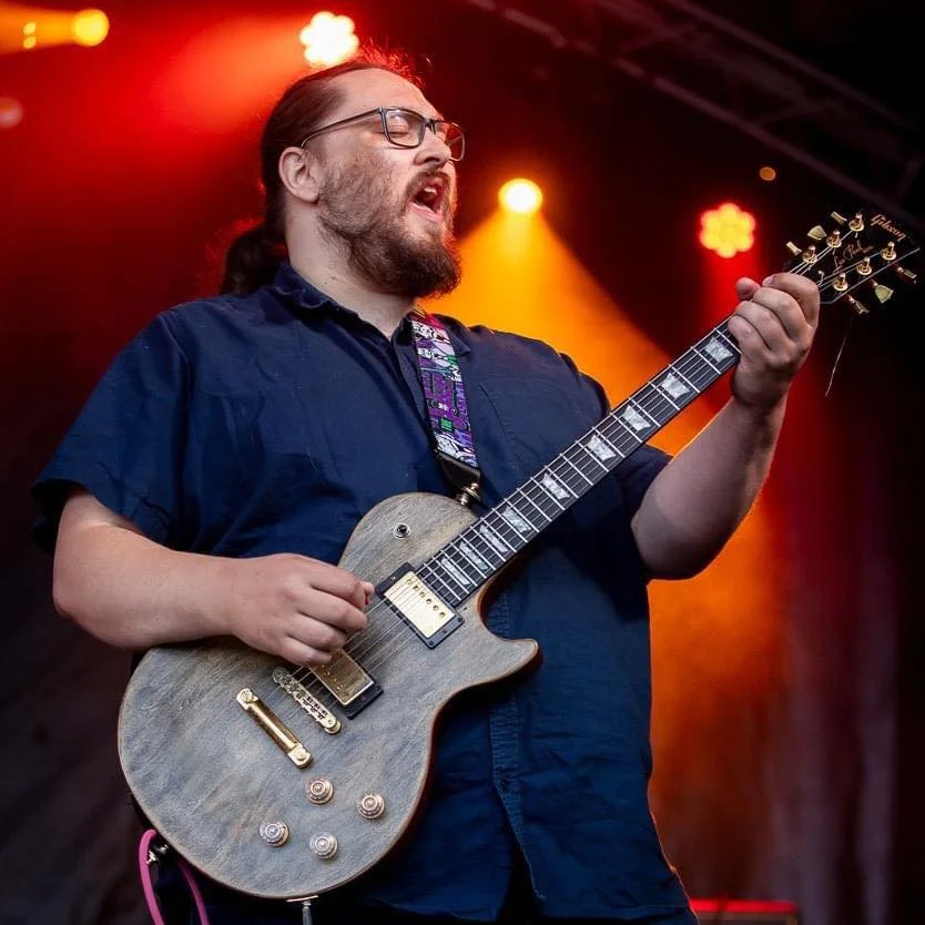 A man with glasses and a beard playing an electric guitar on stage with red and yellow stage lights.