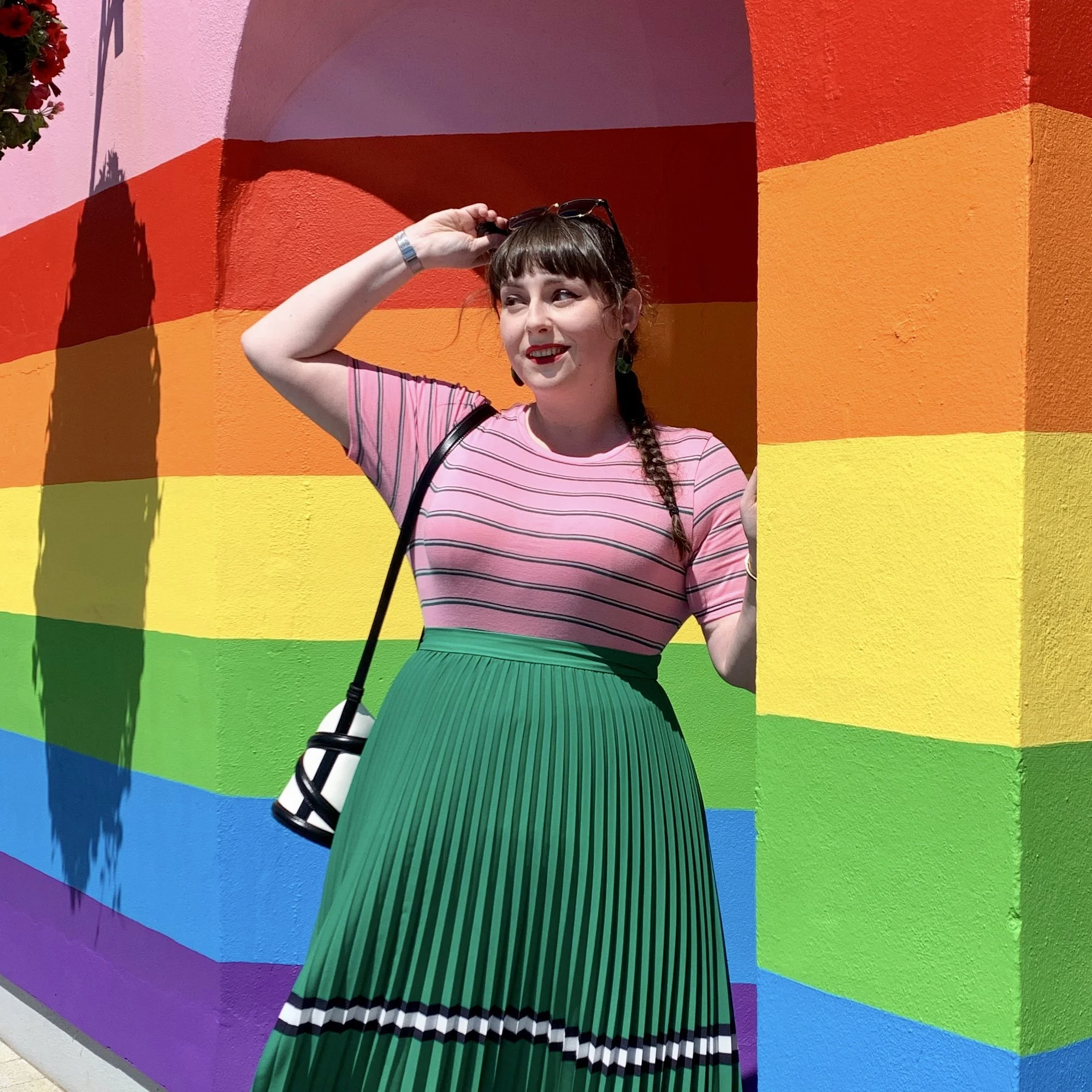 A woman with brown hair styled in braids, wearing a pink striped shirt, green pleated skirt with black and white stripes near the hem, standing in front of a colorful rainbow mural wall.