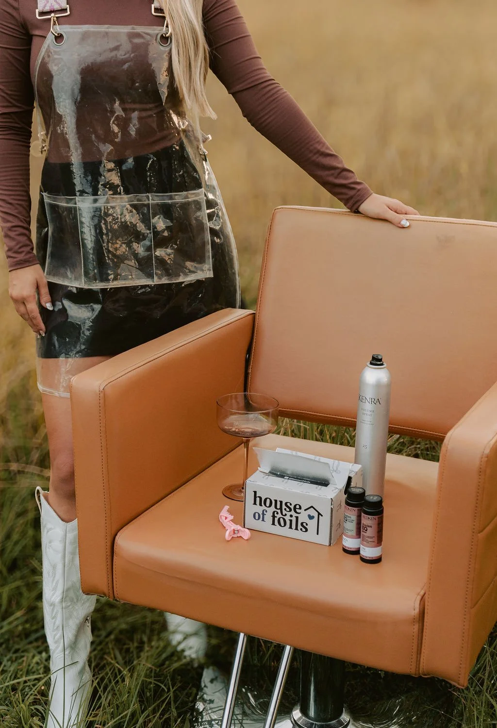 A person in a brown long-sleeve shirt and clear plastic apron standing next to a tan chair outdoors in a field.