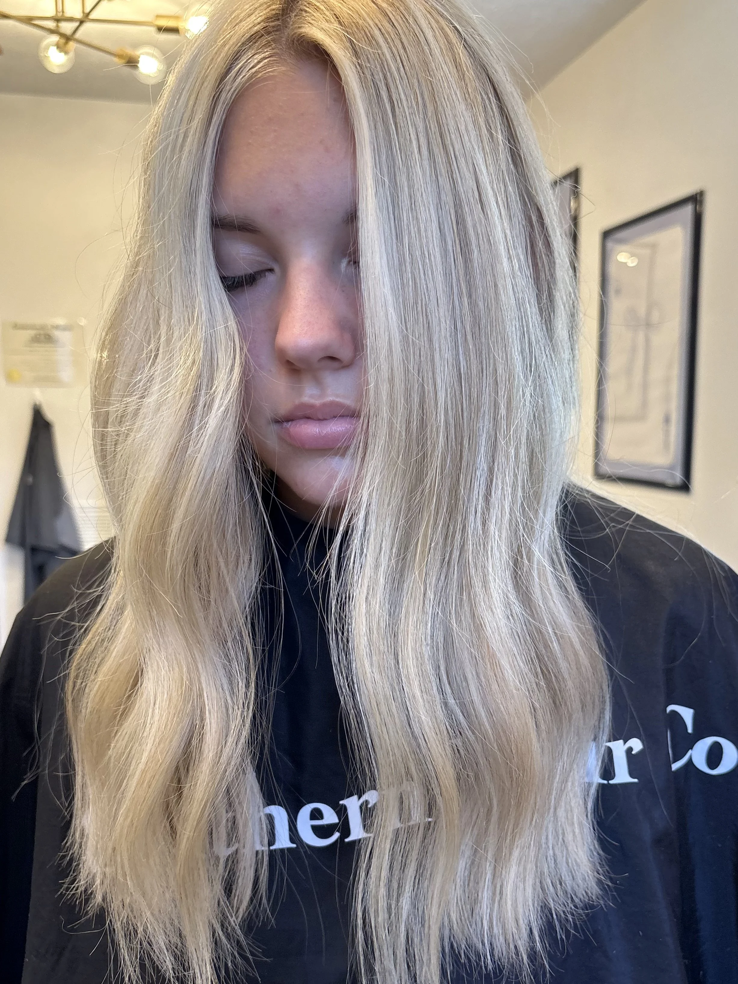 A young woman with long, wavy, blonde hair and fair skin has her eyes closed. She is in a salon or similar setting, wearing a black cape with parts of text visible.