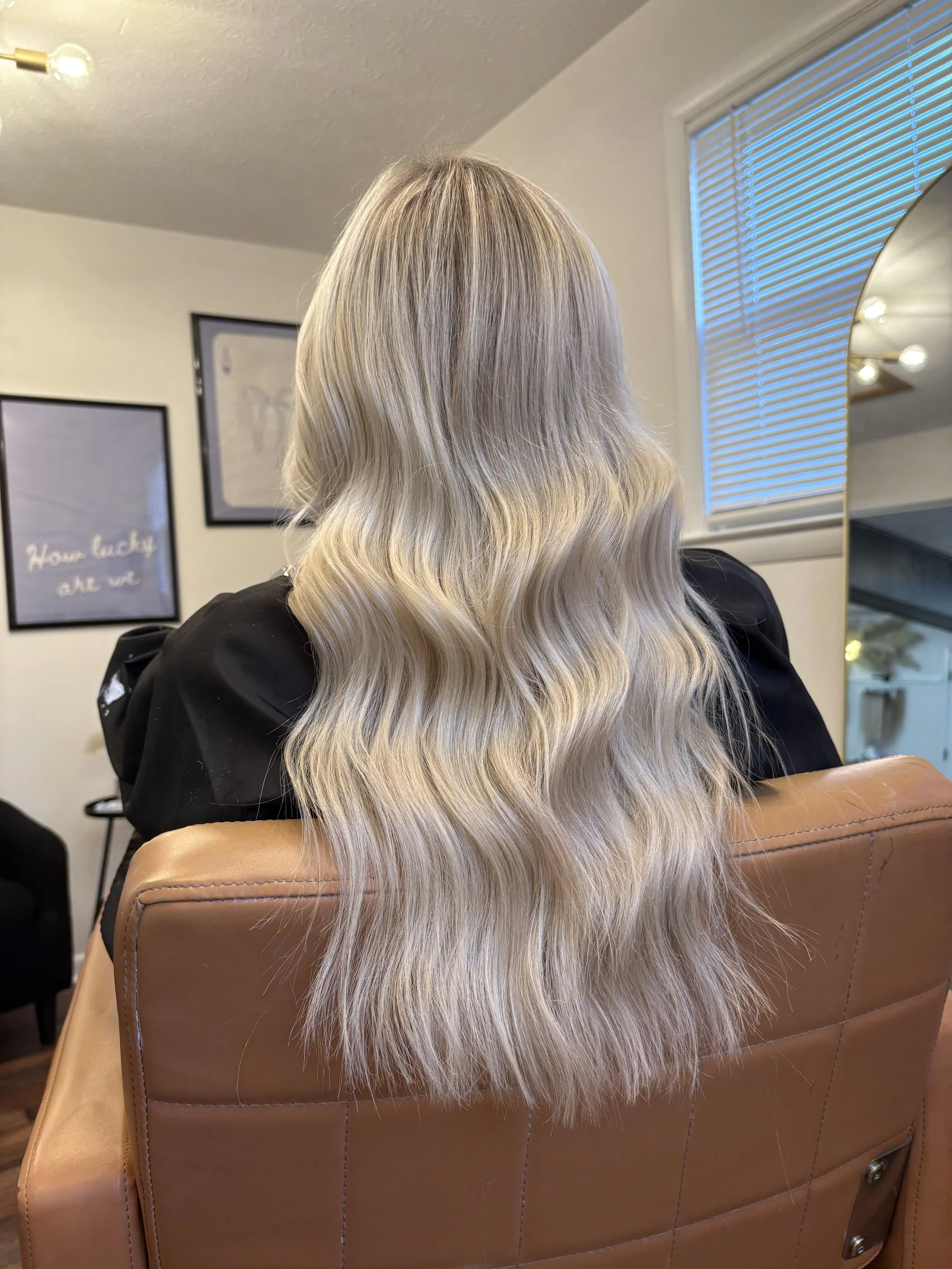 Back of a woman with long, blonde, wavy hair sitting in a salon chair.