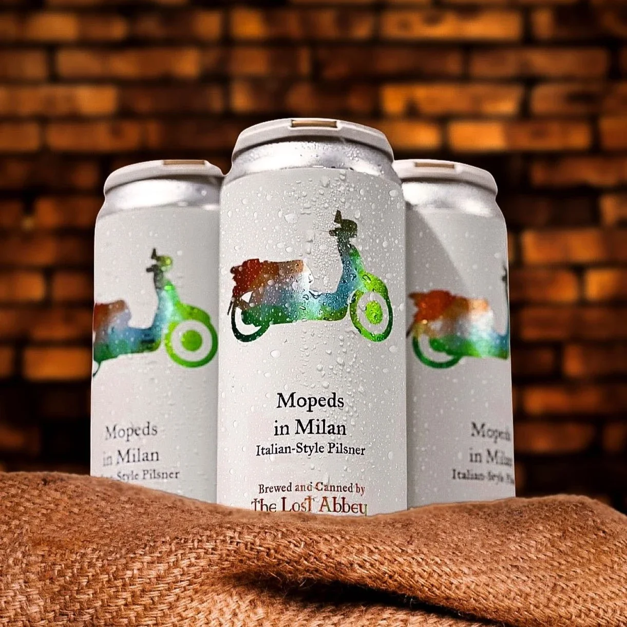 Three cans of Milan-Moped-themed Italian-style pilsner beer with condensation, set against a brick wall background and resting on a textured surface.