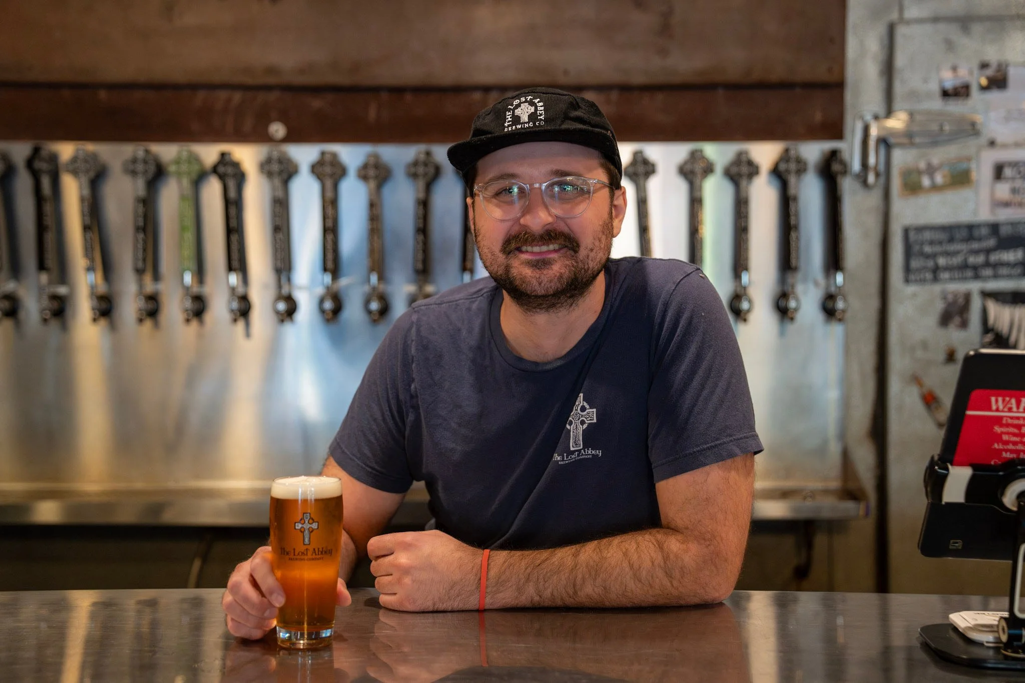 Person in a brown hooded cloak with hands joined in prayer, standing in a dimly lit aisle with bottles on shelves.