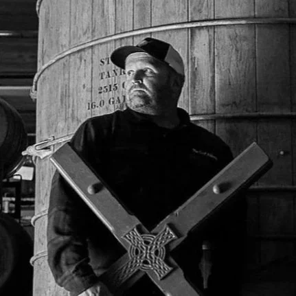 A man wearing a baseball cap and dark clothing standing in front of barrels, holding a graphic resembling swastika symbols.