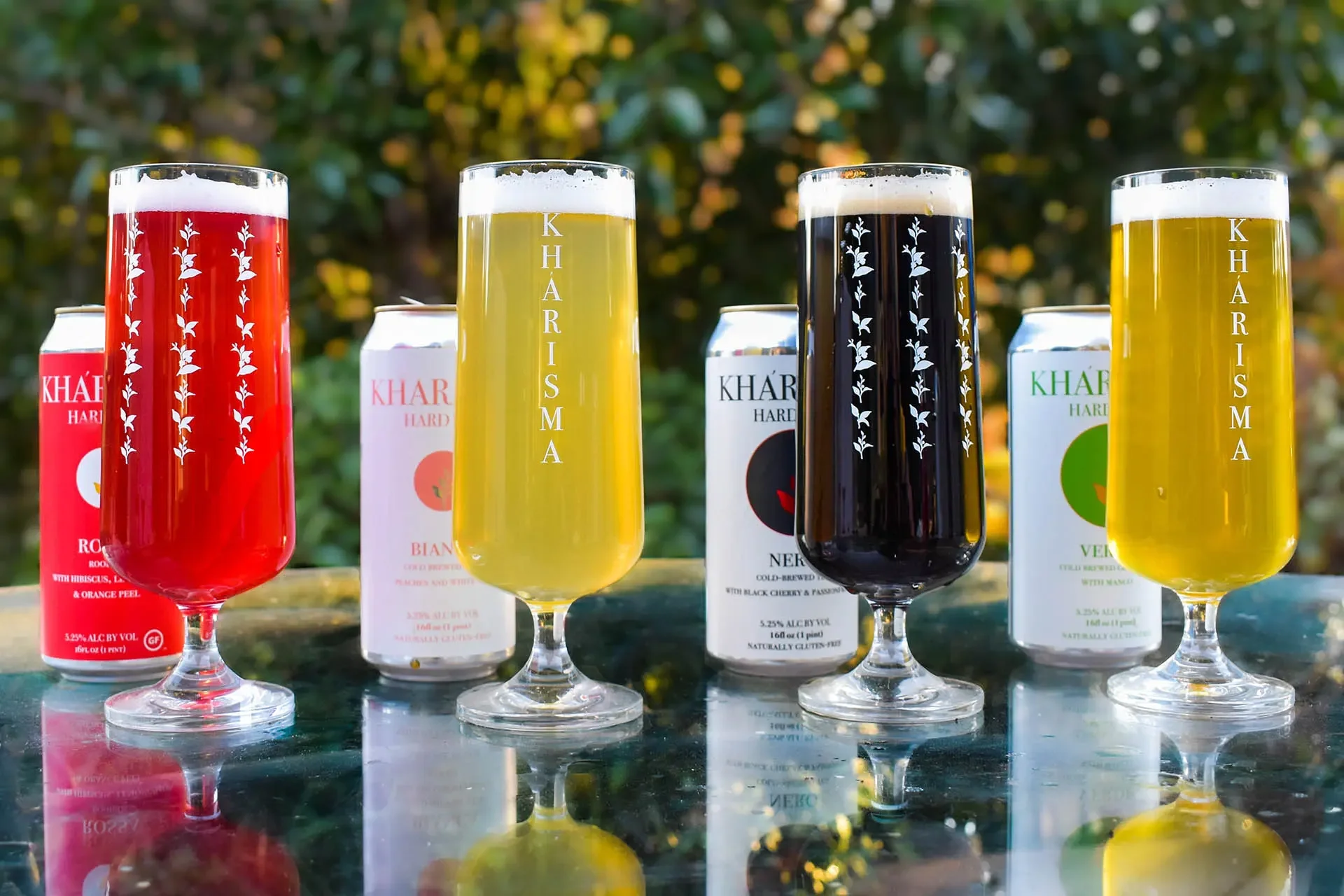 Four tall glasses filled with colorful drinks placed on a reflective surface outdoors with blurred greenery in the background. The glasses have a floral pattern and the brand name 'KHARISMA' printed vertically. Behind each glass is a can or box of beverage with corresponding colors.
