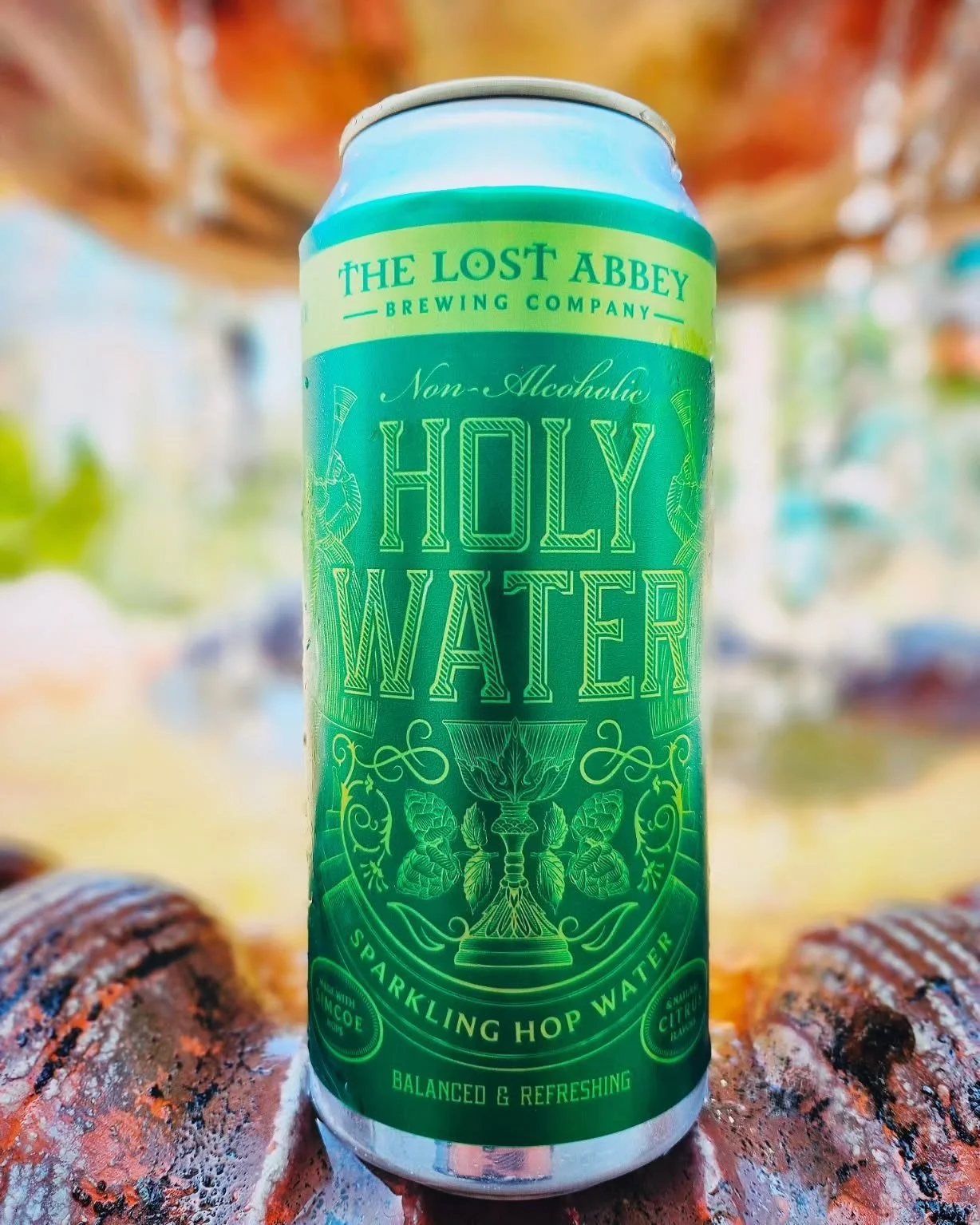 A can of The Lost Abbey Holy Water non-alcoholic sparkling hop water with green label. The can is placed outdoors against a colorful, blurred background.