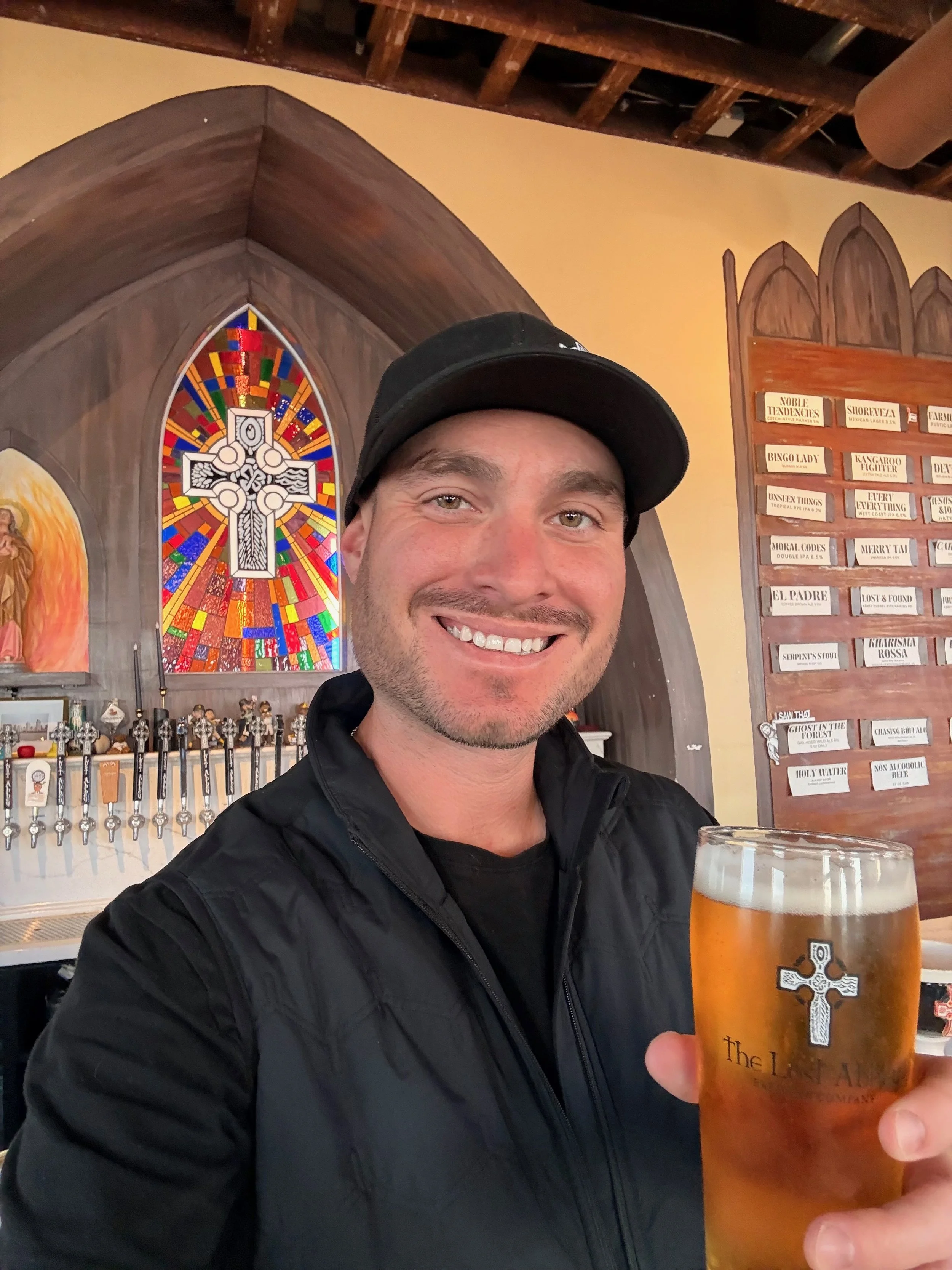 Person in a hooded cloak with hands in prayer position in a dimly lit wine cellar or storage room with shelves of bottles.