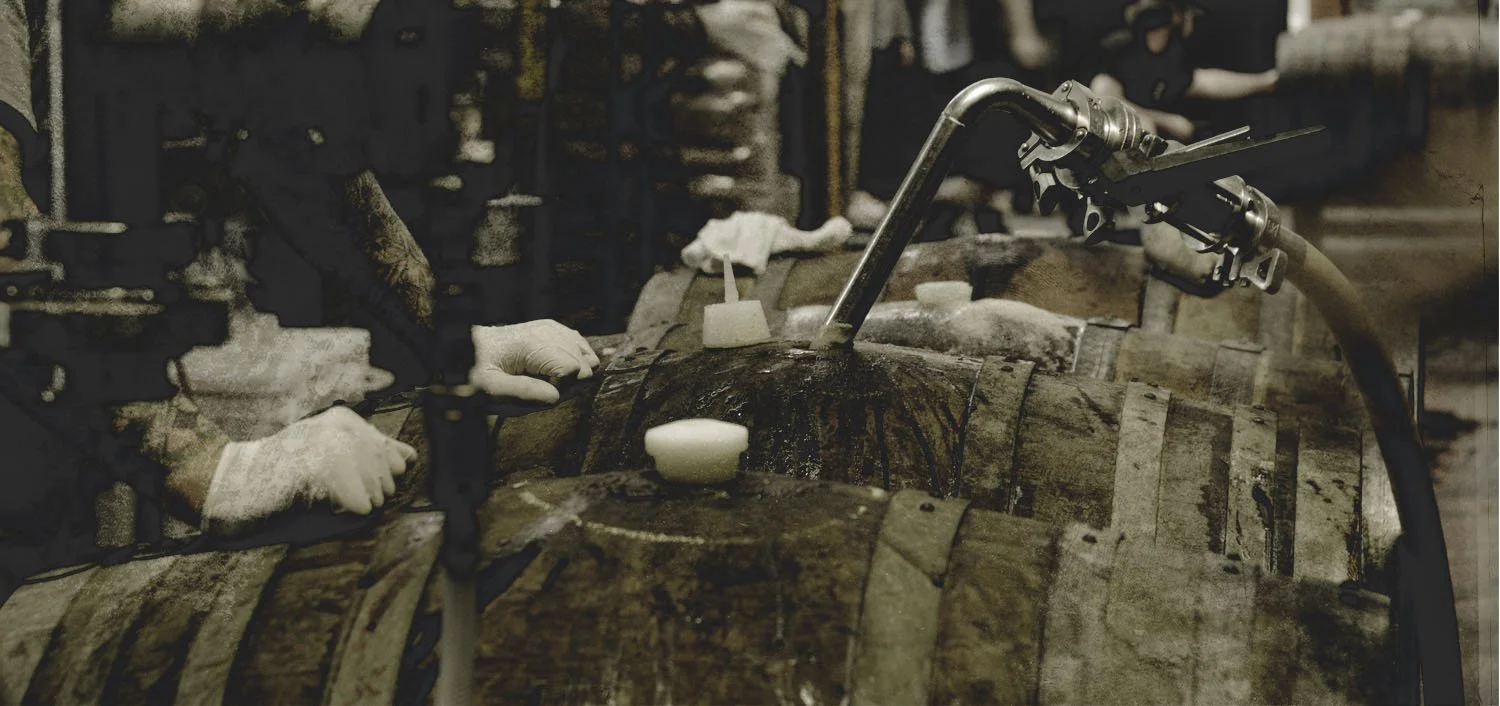 People working with barrels of wood, likely involved in a wine or whiskey production process, with one person wearing gloves.