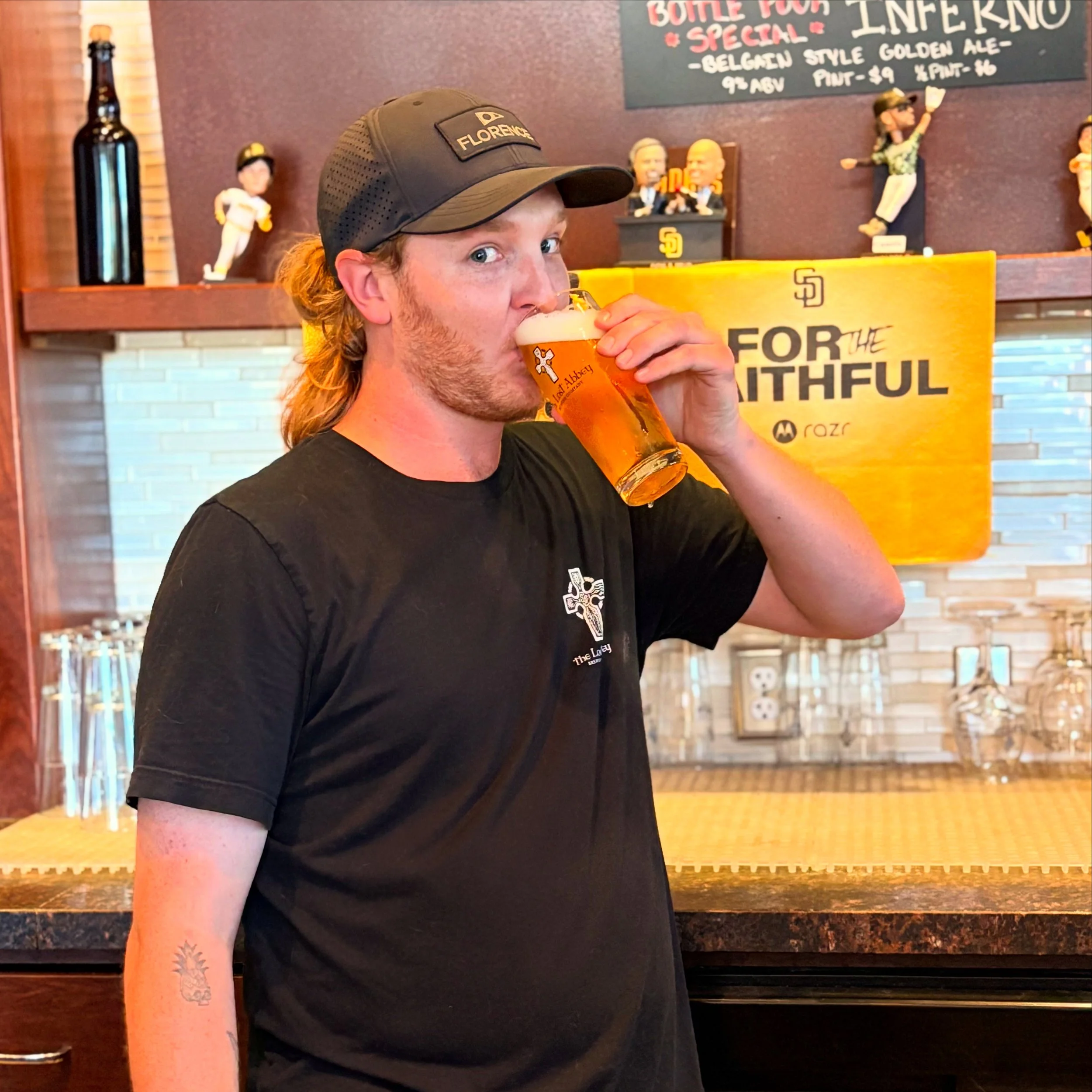 A man with long hair and a beard wearing a black t-shirt and a black baseball cap is drinking beer from a clear glass at a bar.